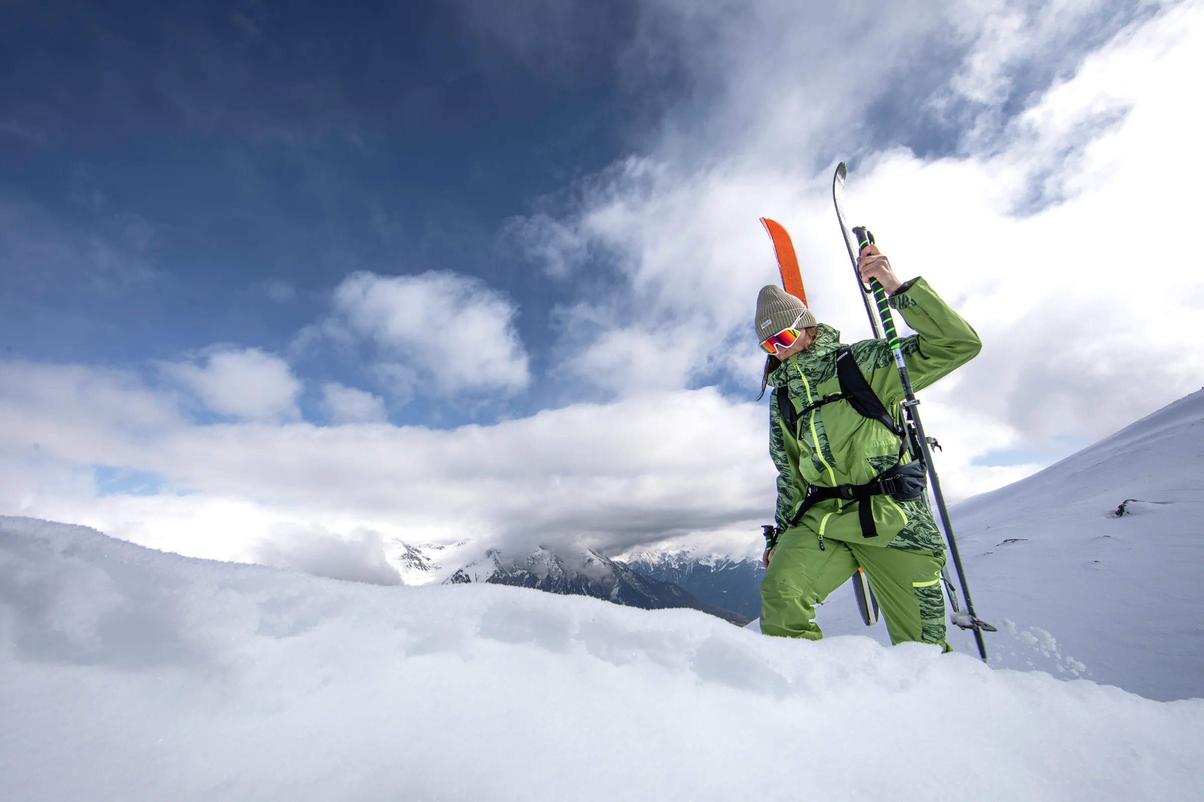 Martina Wengenmeir steigt mit Ski am Rucksack und Stöcken durch tiefen Schnee, hinter ihr verschneite Berge unter blauem Himmel und Wolken.