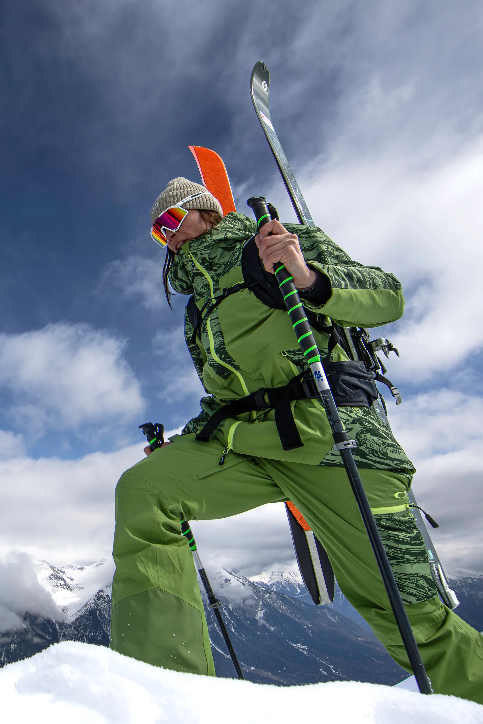 Martina Wengenmeir steigt in grüner Skikleidung mit Ski am Rucksack und Stöcken durch den Schnee, aus tiefer Perspektive vor Bergen und Wolken gesehen.