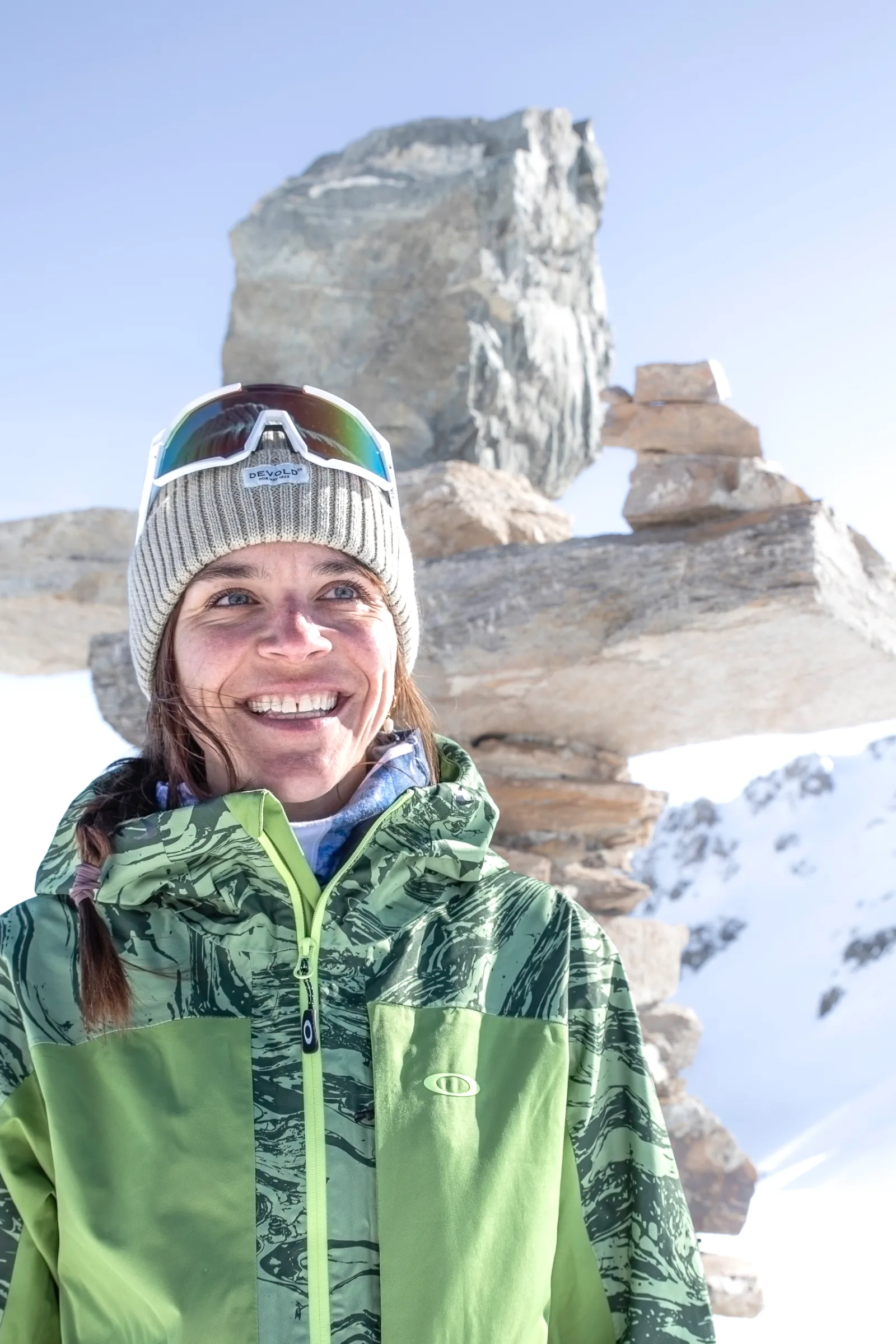 Martina Wengenmeir lächelt in grüner Jacke und grauer Strickmütze mit hochgeschobener Sonnenbrille vor aufgeschichteten Steinen und verschneiten Bergen.