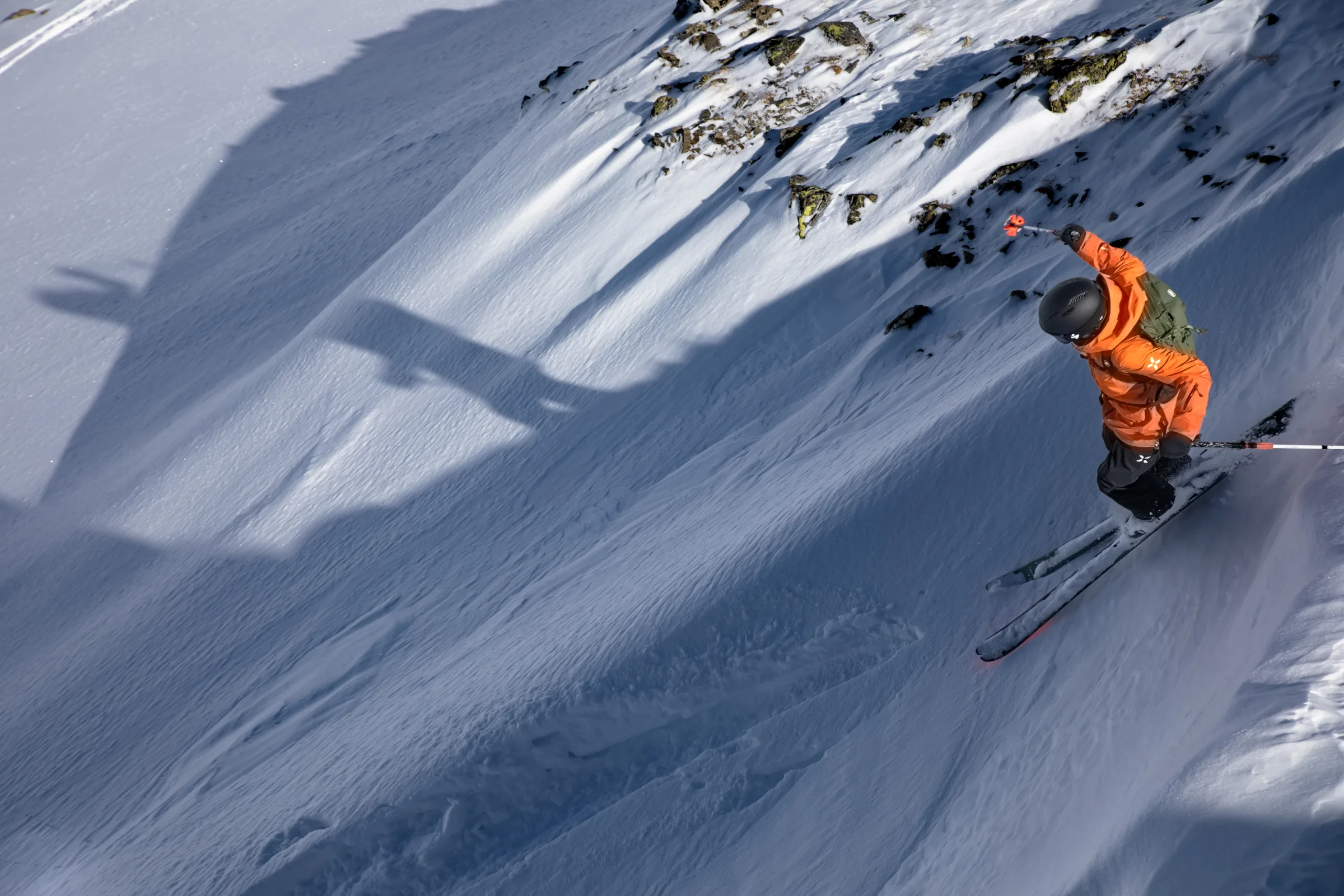Martina Wengenmeir fährt in orangefarbener Jacke mit schwarzem Helm und grünem Rucksack einen steilen Schneehang unterhalb freiliegender Felsen hinab.