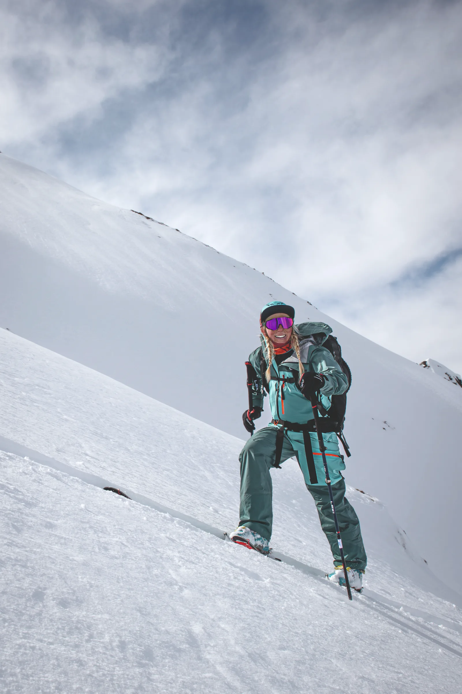 Michaela Casotti smiles with skis, poles and a backpack on a steep snowy slope in Gastein, with a snow-covered ridge behind her.