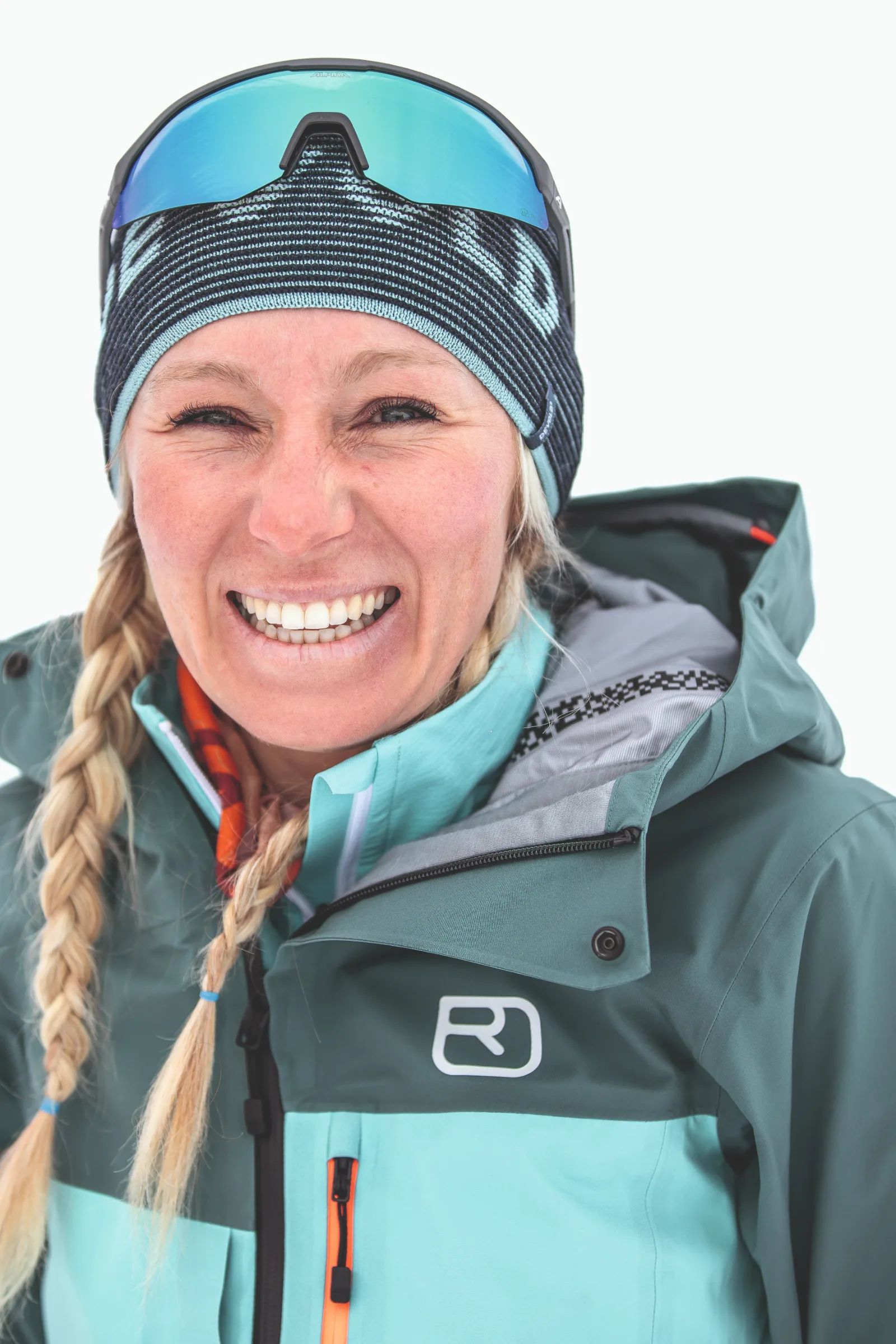 Michaela Casotti smiles in close-up against a white background, with blonde braids, a striped beanie and mirrored sunglasses resting on top.