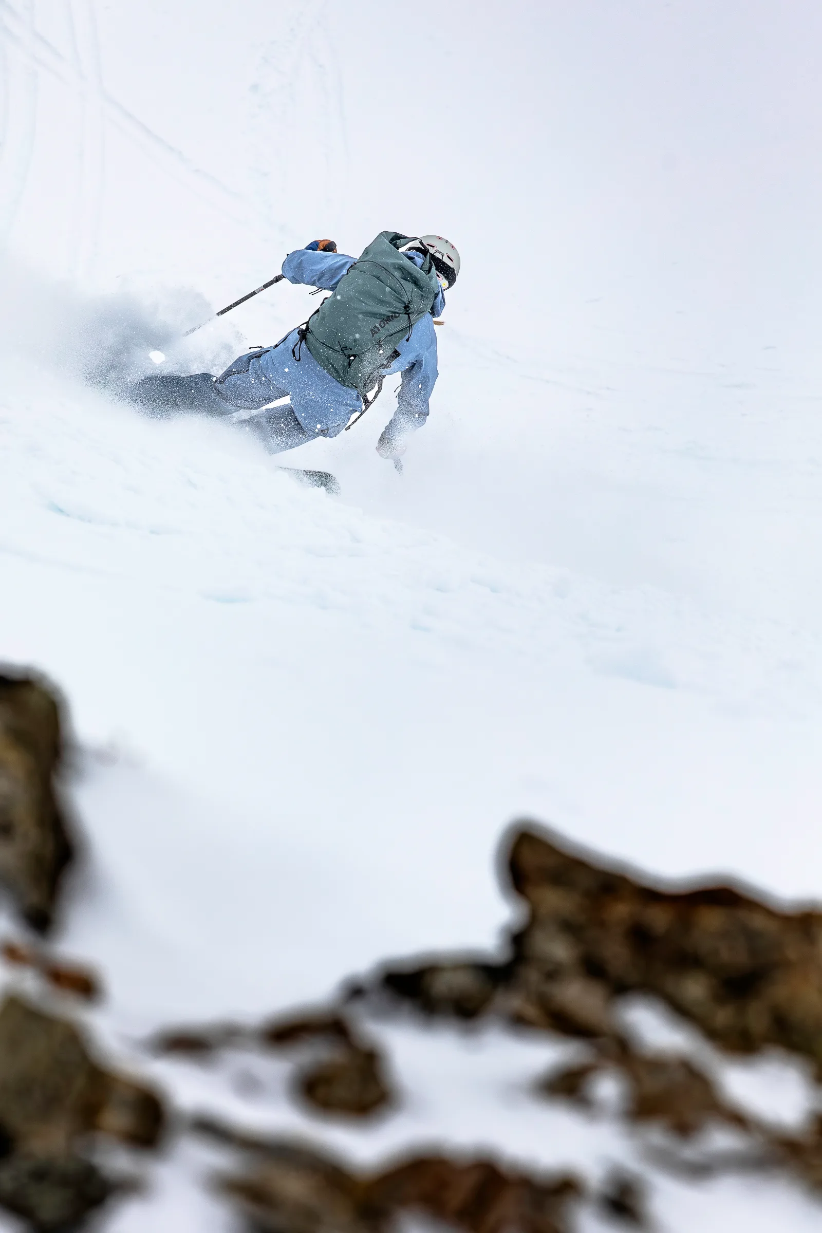 Seen from behind, Michaela Casotti skis through a spray of snow with a greenish backpack, while blurred rocks frame the slope in the foreground.