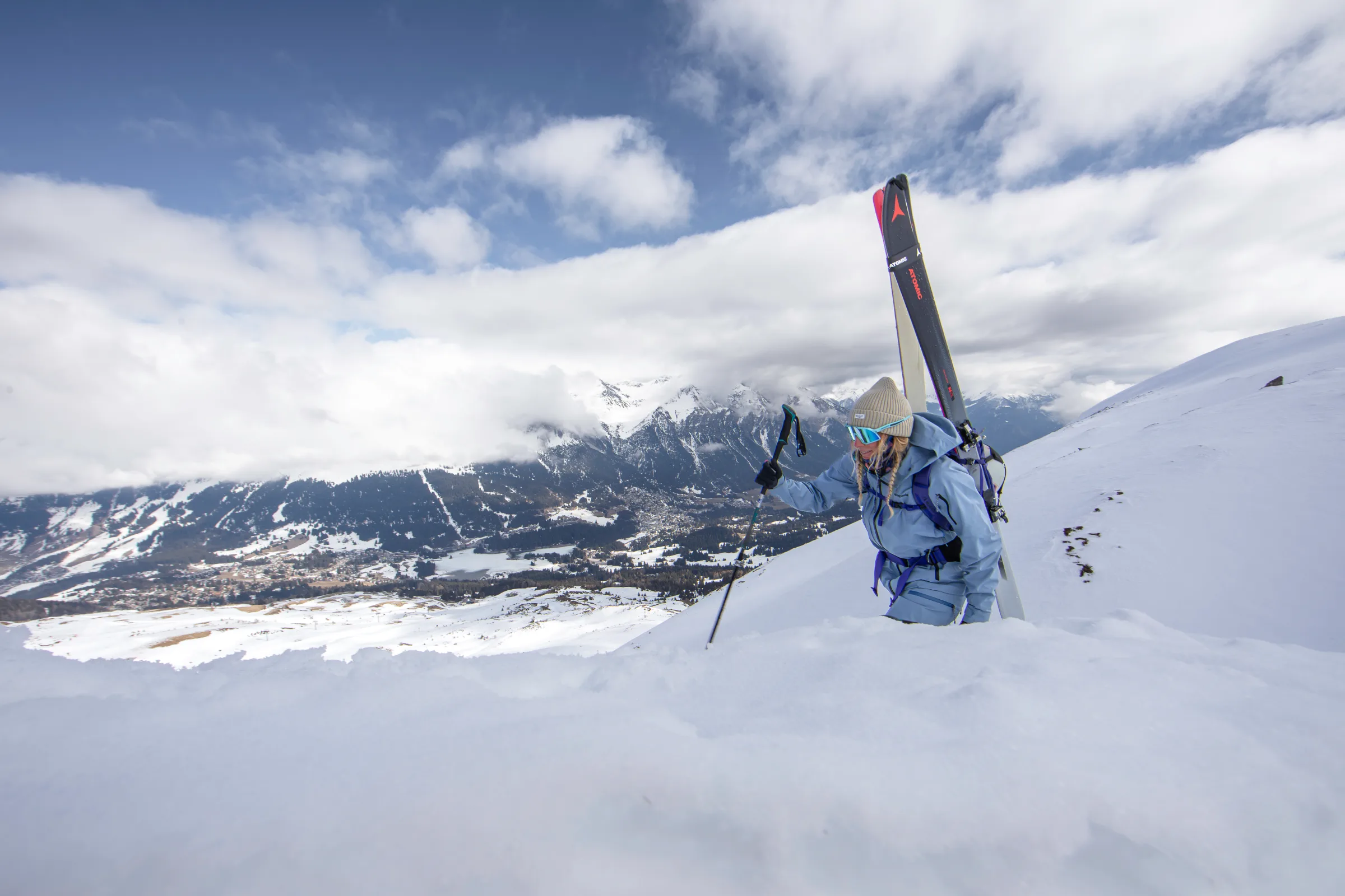 Michaela Casotti climbs a snowy slope above Lenzerheide with skis on her backpack and a pole, overlooking the valley and cloud-covered mountains.