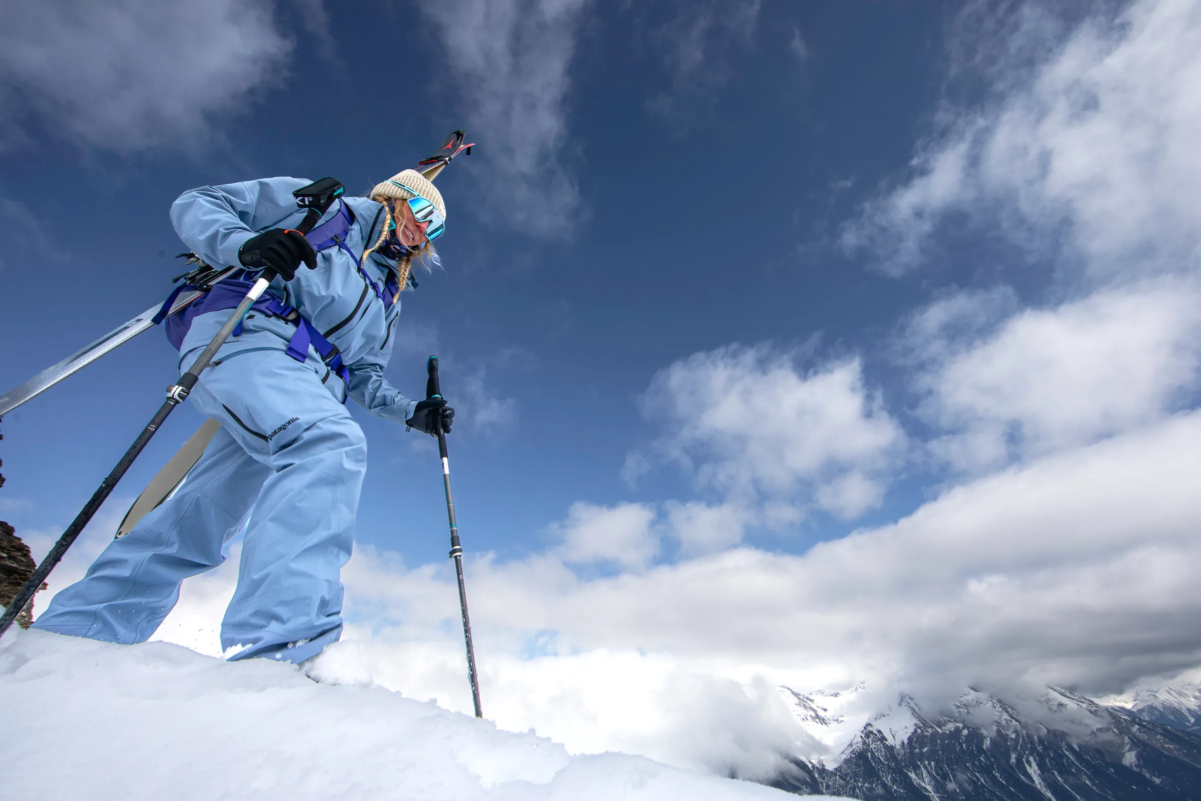 Viewed from below, Michaela Casotti climbs a snowy slope with skis on her backpack and two poles beneath blue sky and white clouds.