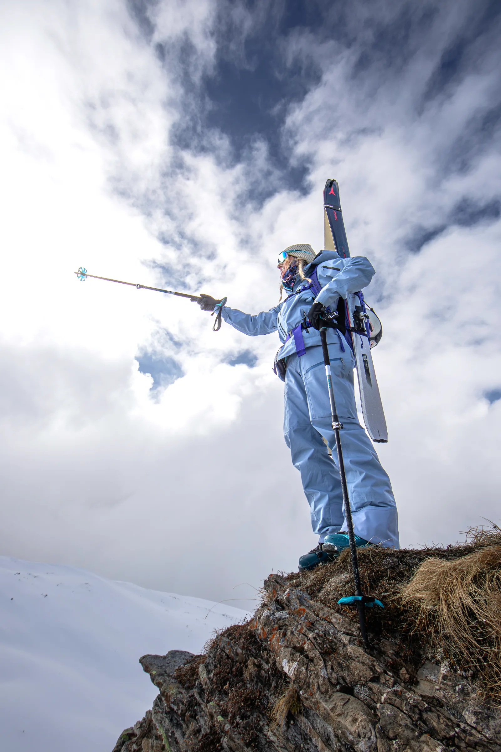 Michaela Casotti stands on a grassy rock ledge with skis on her backpack, extending one pole to the left beneath a cloudy sky.