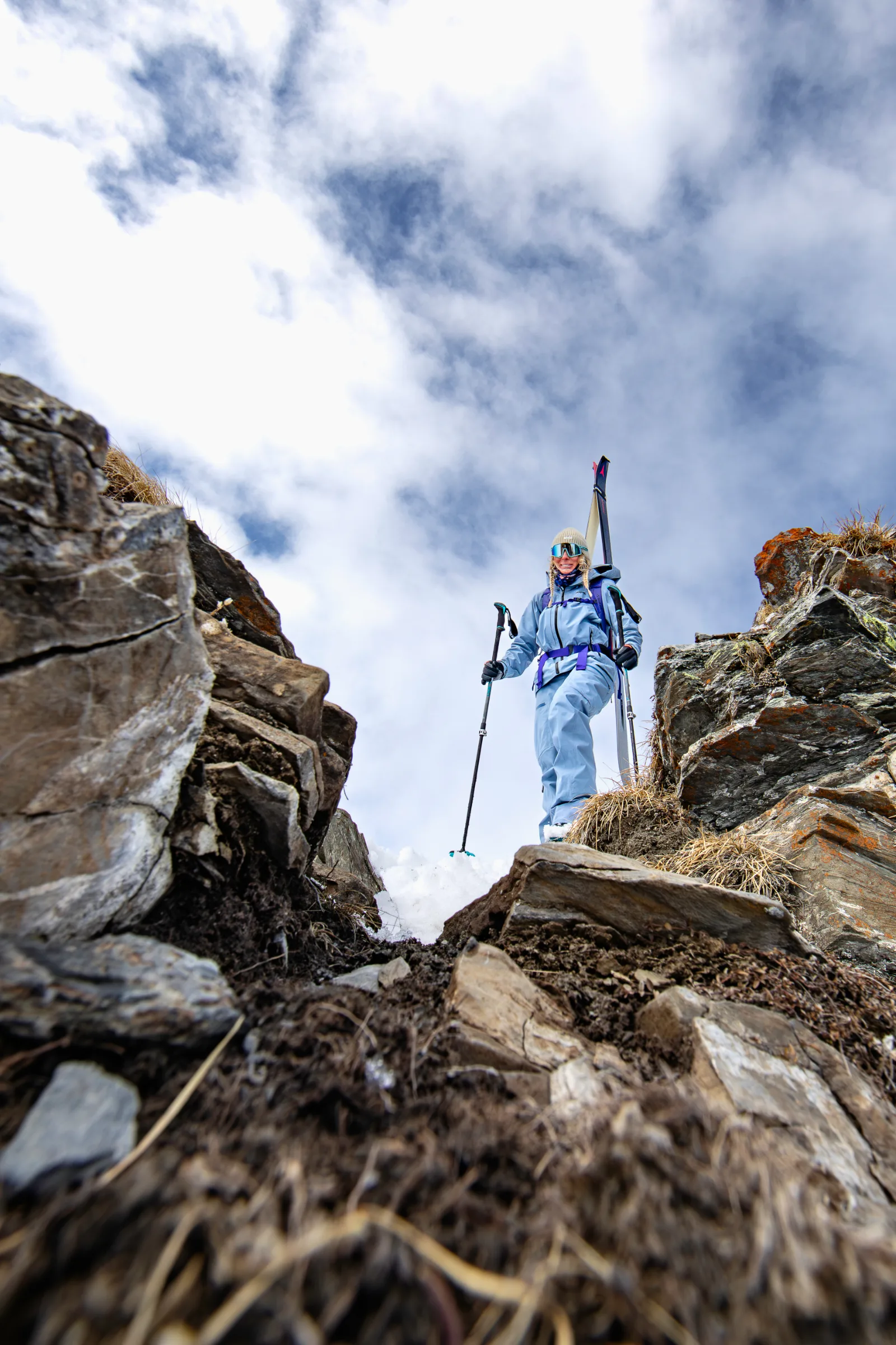Michaela Casotti stands in light blue ski clothing with skis on her backpack and poles in hand between snowy rocks beneath a cloudy sky.