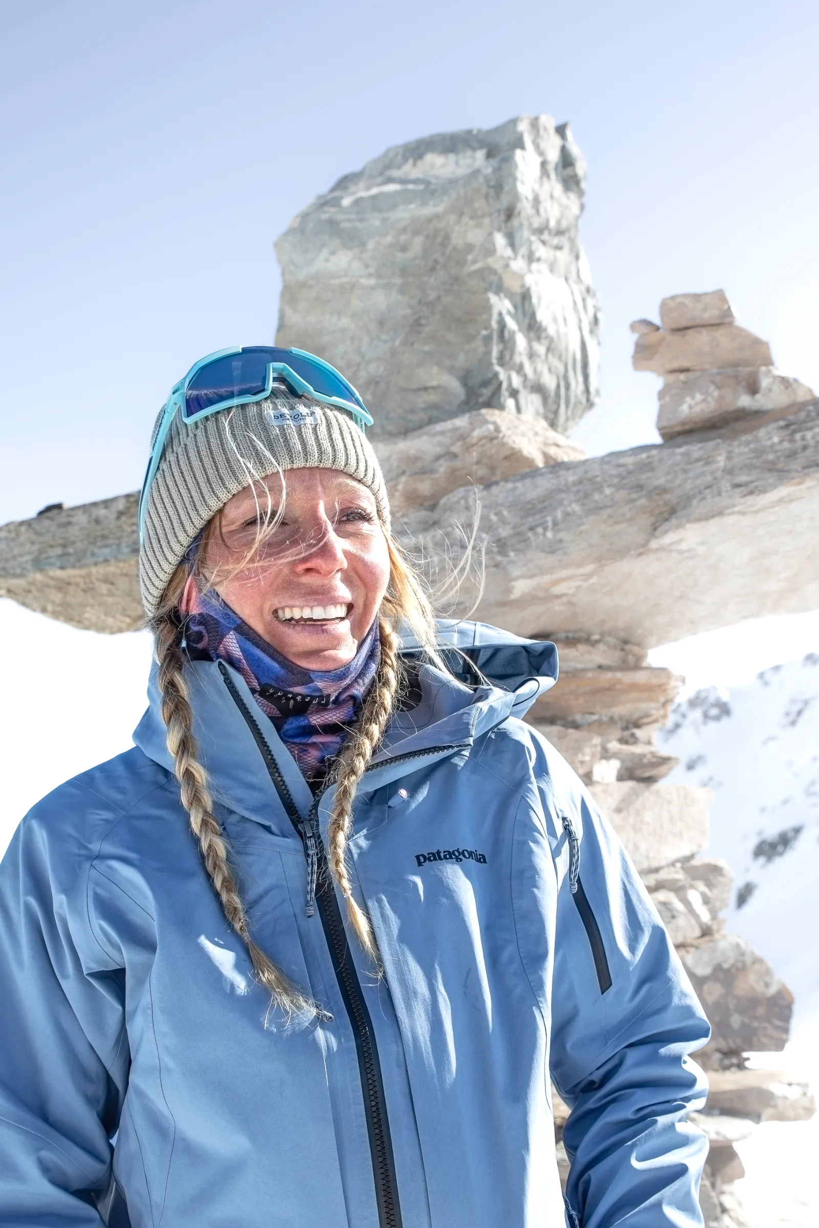 Michaela Casotti smiles with braided hair, a knitted beanie and goggles pushed up on her hat, with stacked stone slabs and snow behind her.