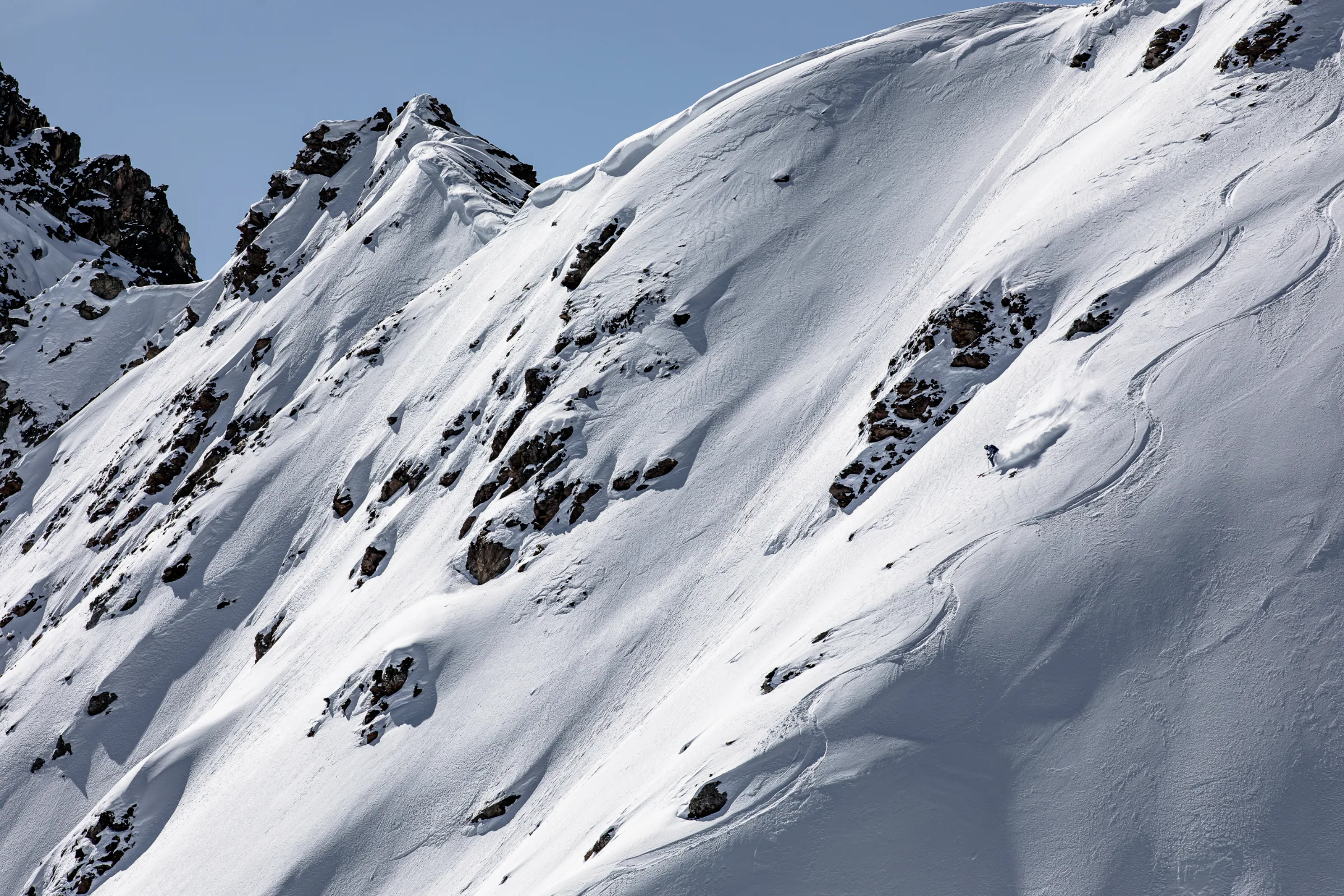 Michaela Casotti skis down the steep snowy slope below a cluster of rocks, trailing a cloud of powder.