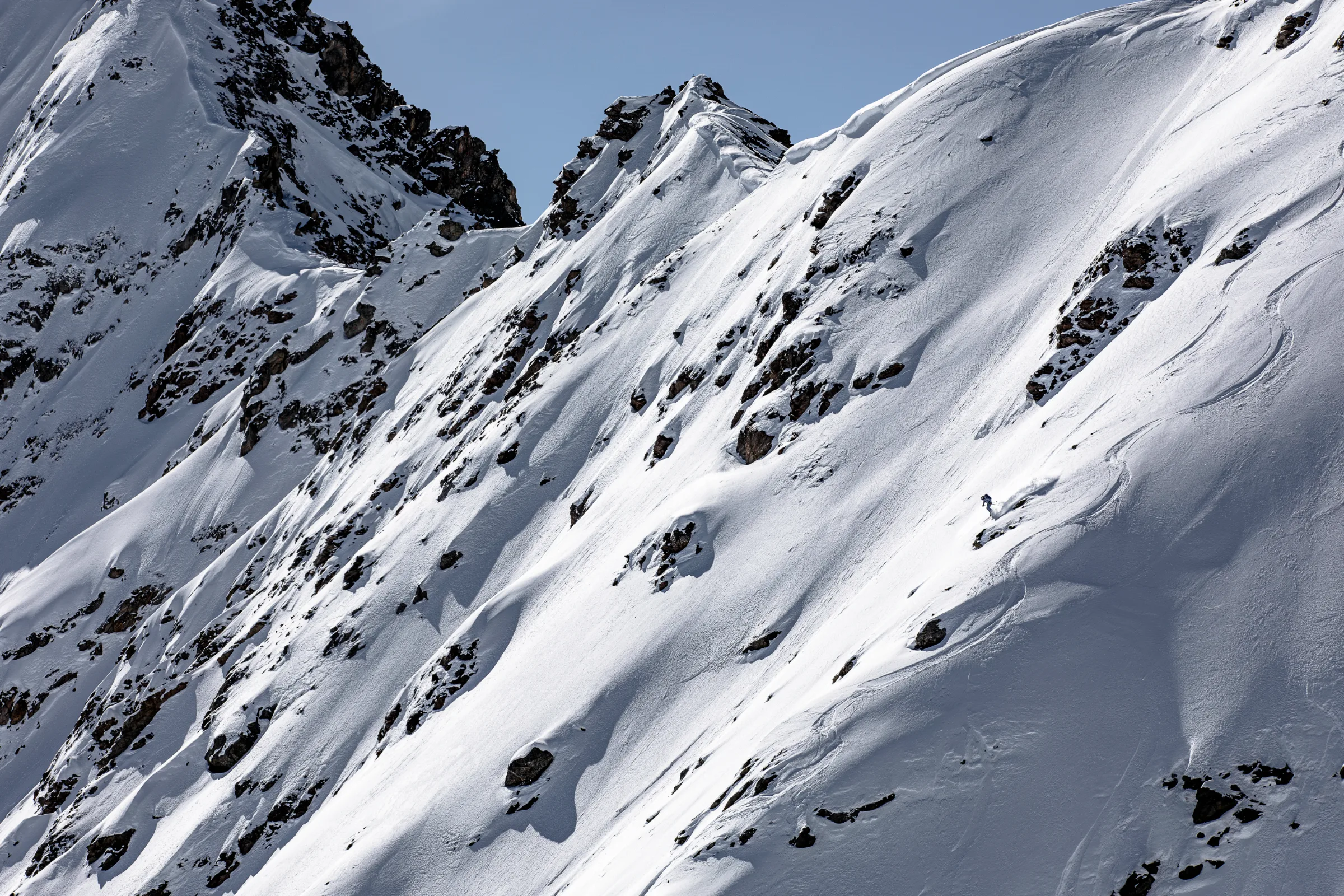Michaela Casotti skis between exposed rocks on a broad snowy mountainside beneath jagged ridges.