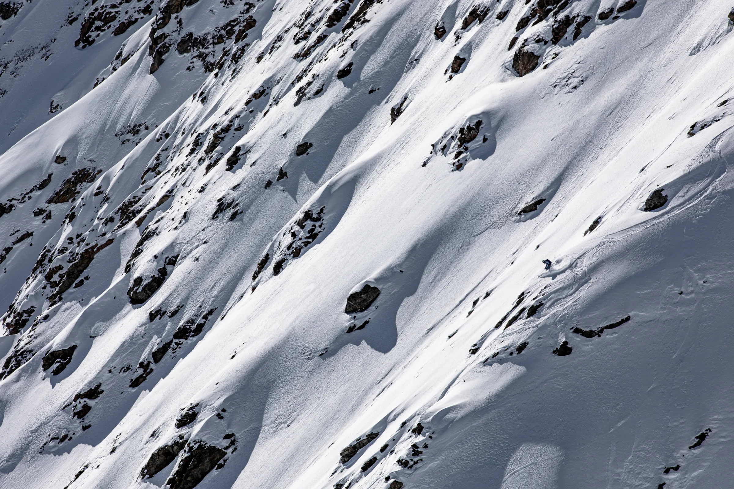 Michaela Casotti leans forward while skiing down a steep, rock-studded snow slope, kicking up snow.