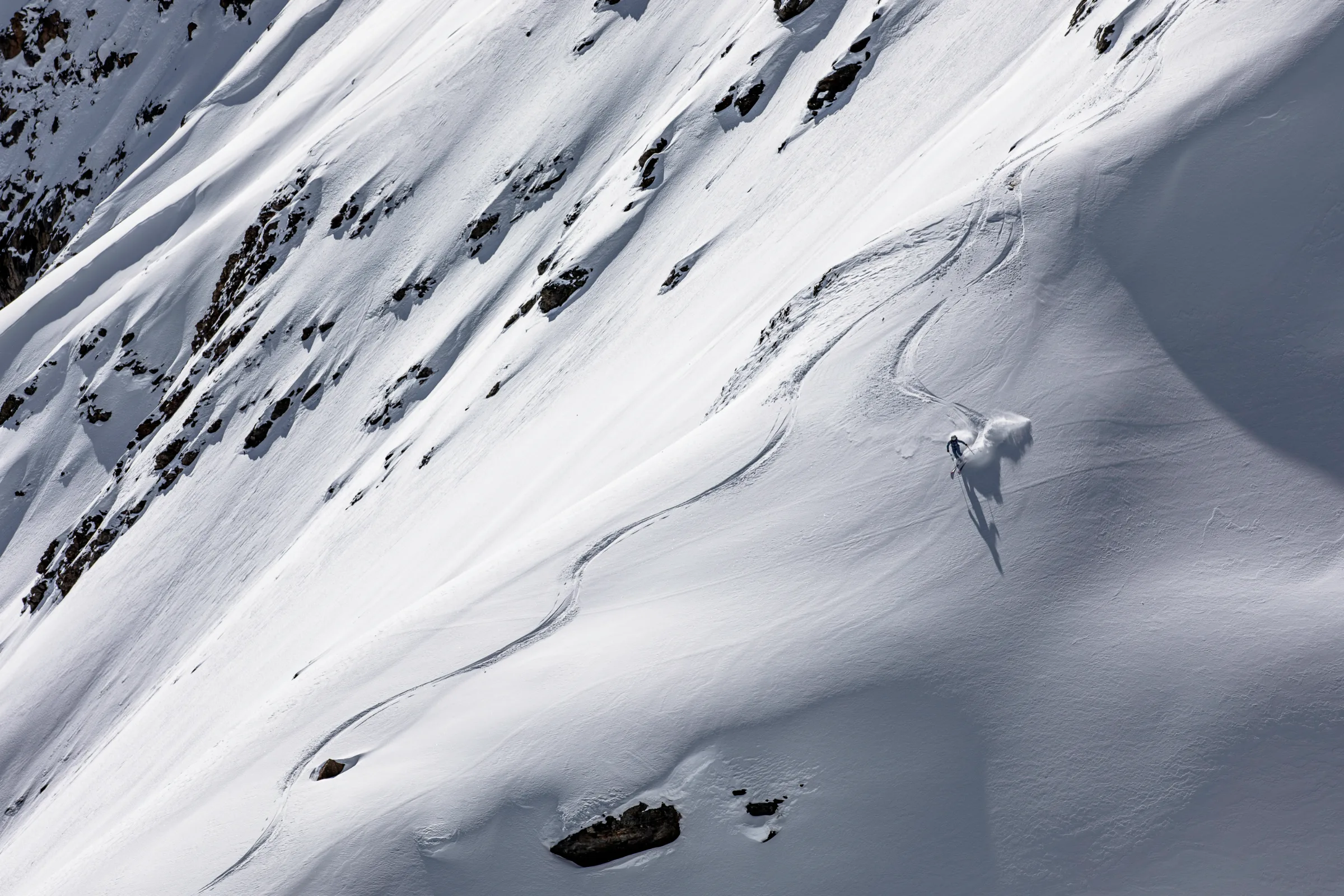 Michaela Casotti turns across an open snow slope, with a tall plume of powder behind her and a long shadow stretching downhill.