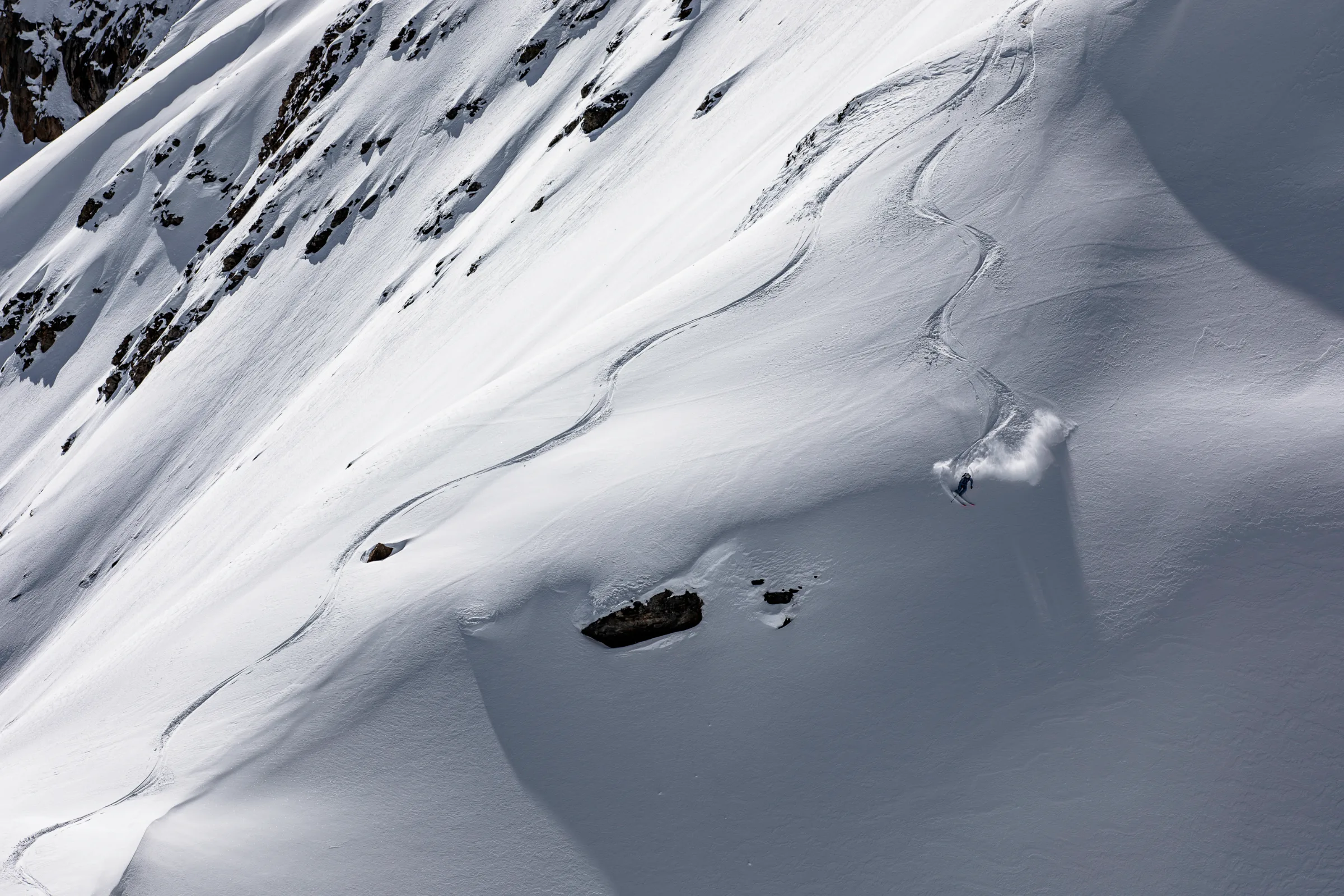 Michaela Casotti makes a sweeping turn above exposed rocks, leaving a curved track and a spray of powder.