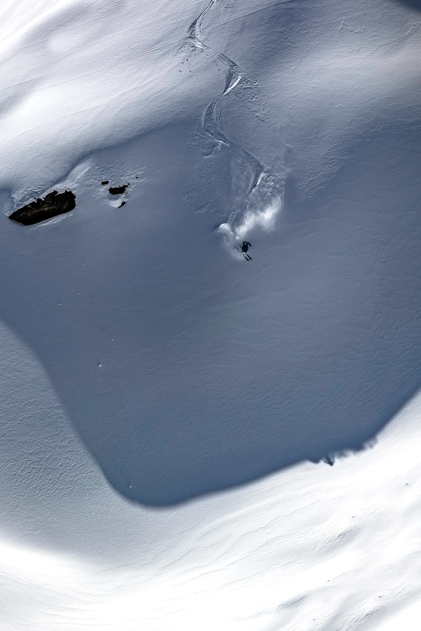 Michaela Casotti skis through a shaded snow bowl below curving ski tracks, with powder billowing behind her and rocks to the left.