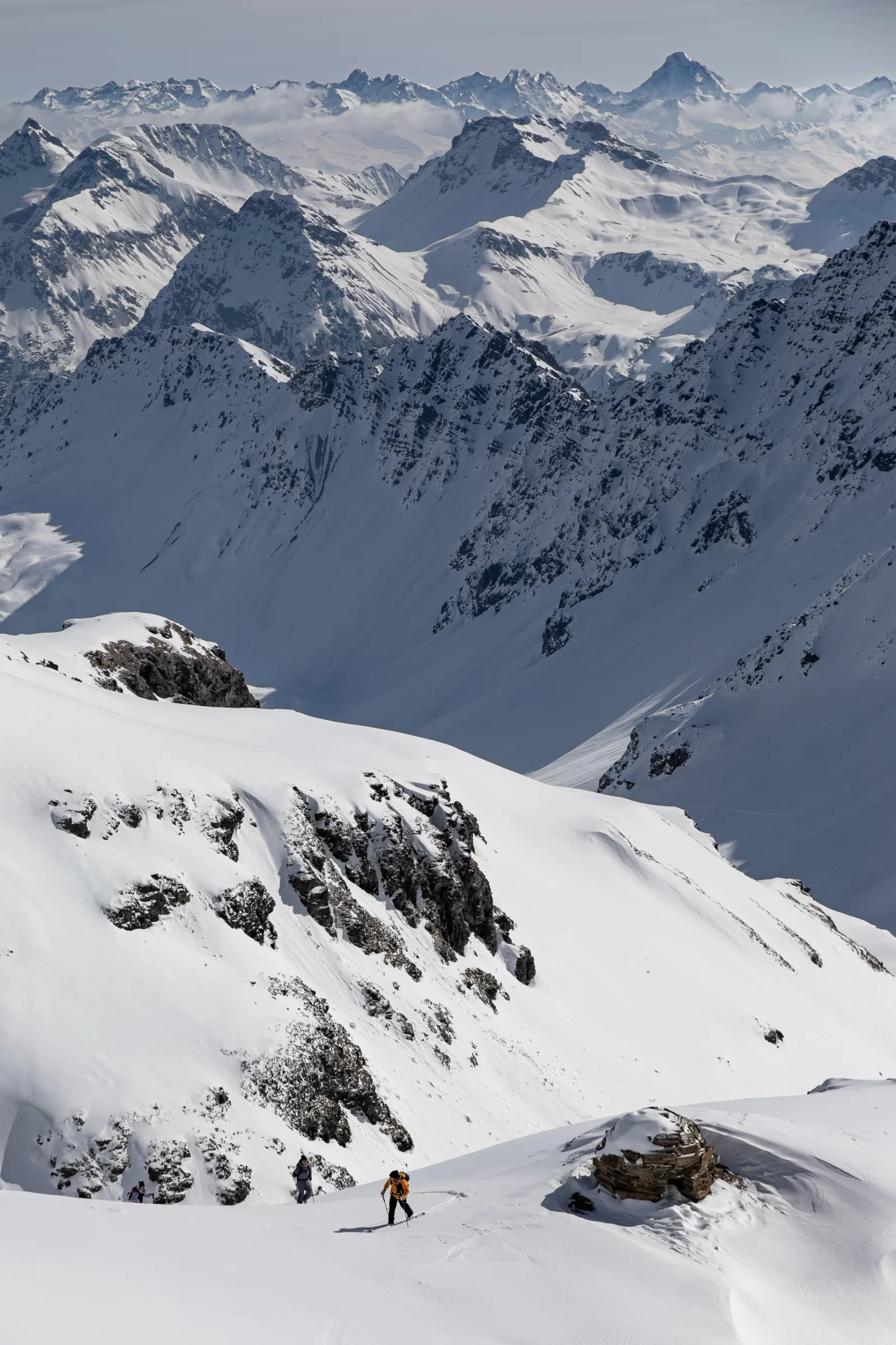 Three ski tourers climb a snowy slope near Lenzerheide beneath a sweeping panorama of snow-covered rocky peaks.