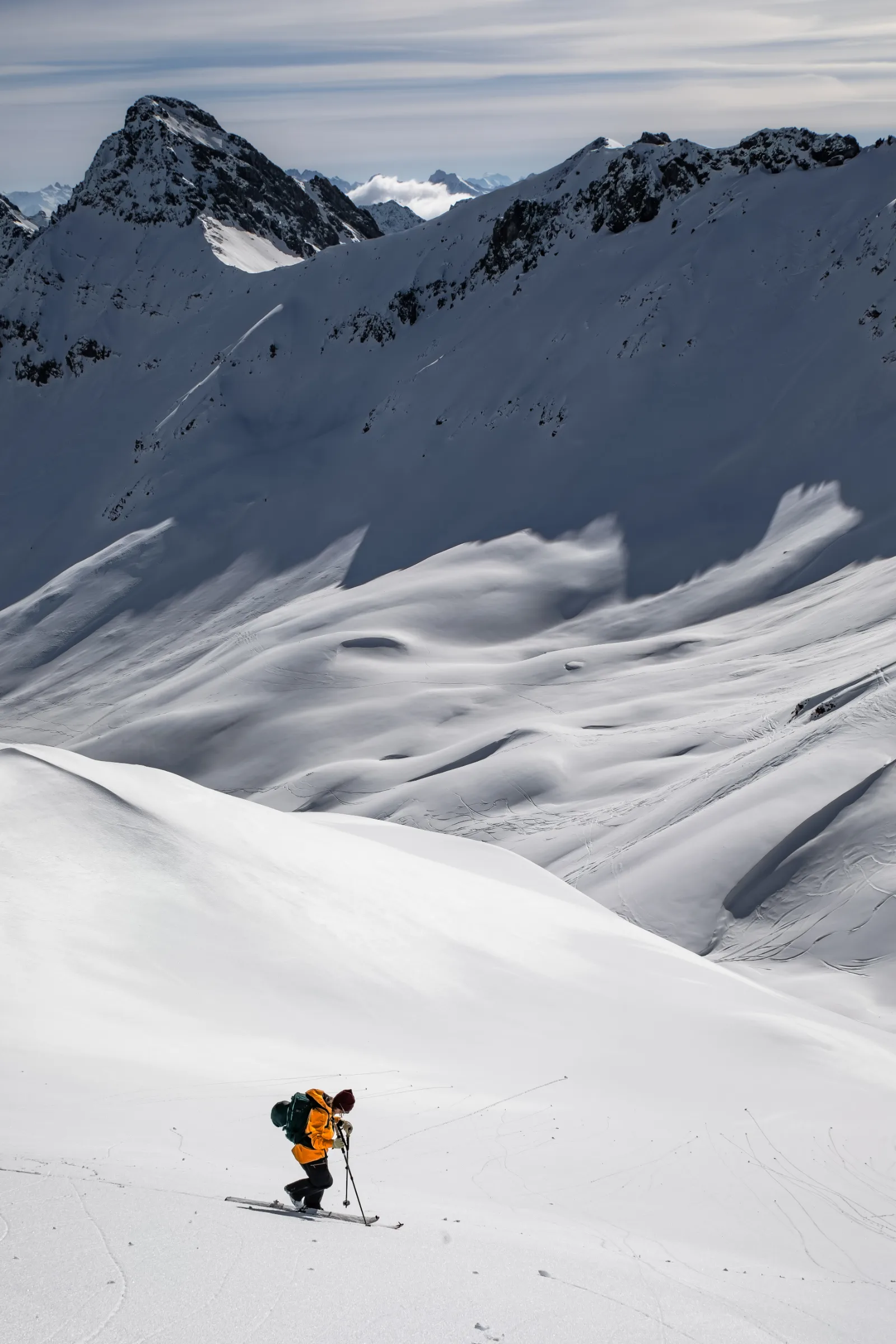 Michaela Casotti climbs on touring skis in an orange jacket and green backpack, with a broad snowy basin and rugged peaks behind her.
