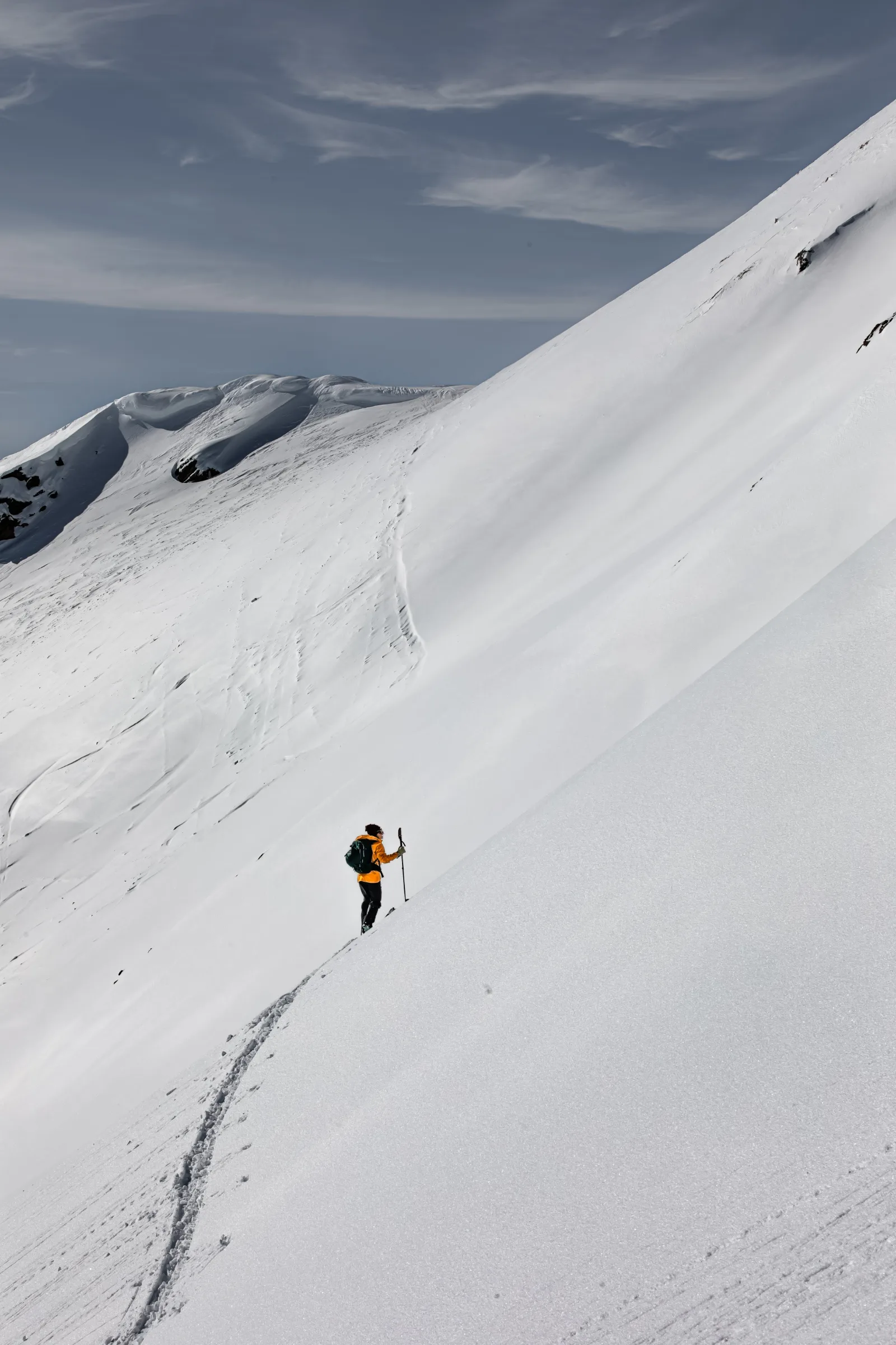 Michaela Casotti follows a narrow climbing track across a steep, open snow slope with touring skis and poles.