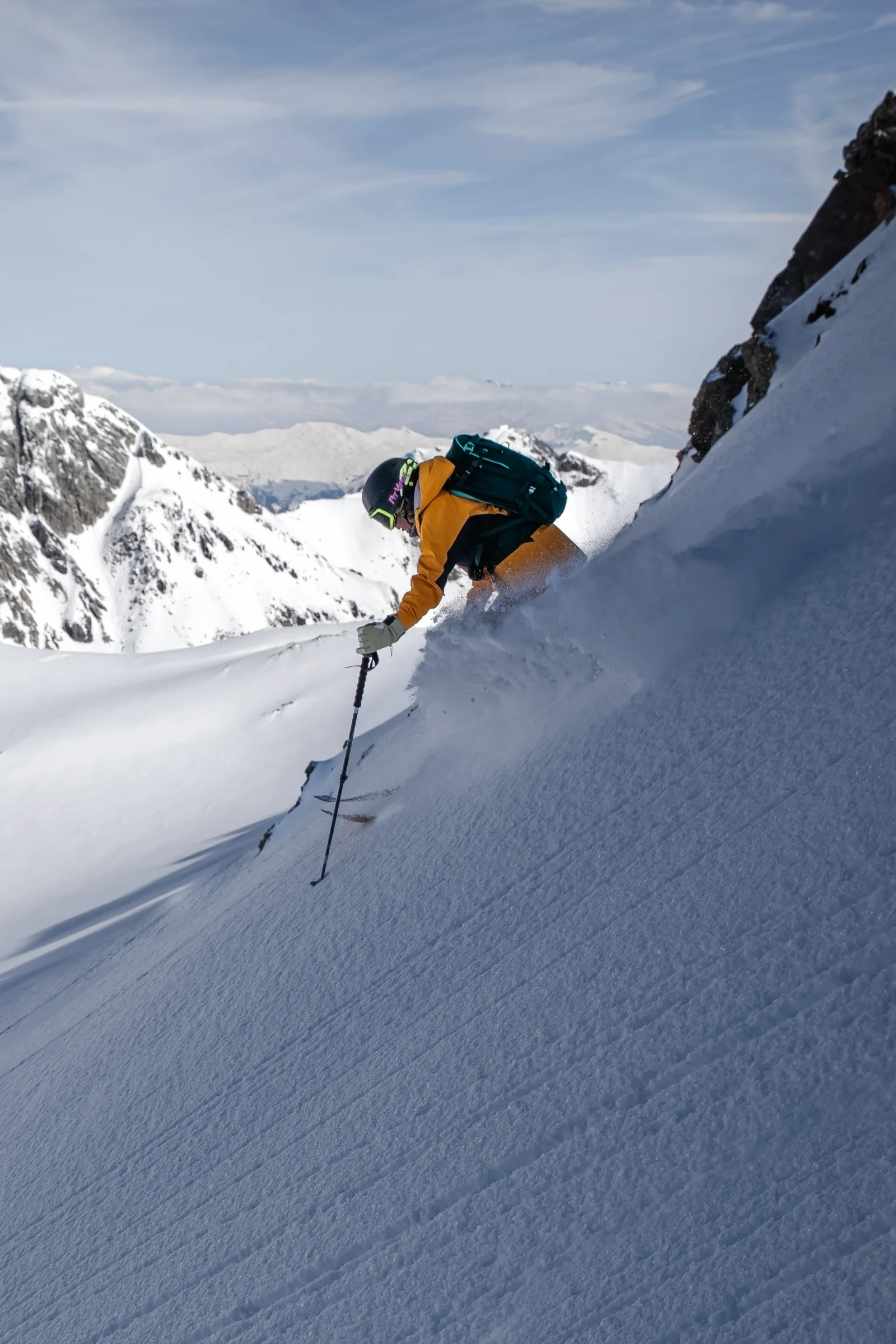 Michaela Casotti crouches low while skiing a steep slope beside a rock face, kicking up snow with snowy peaks behind her.