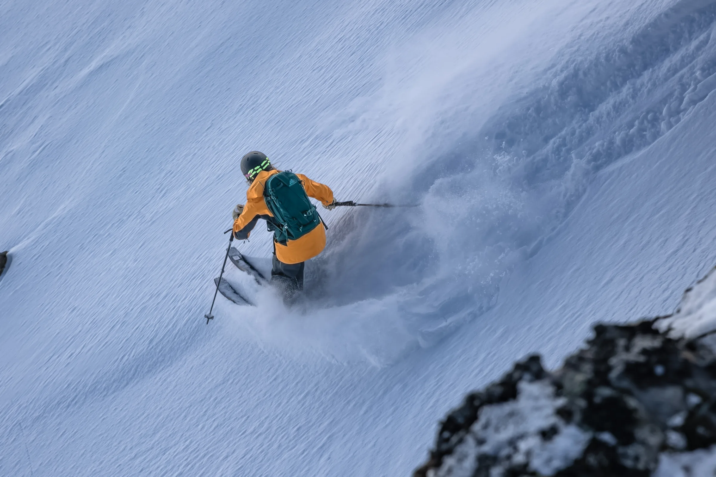 Seen from behind, Michaela Casotti makes a tight turn on a steep slope, sending a plume of snow up beside her green backpack.