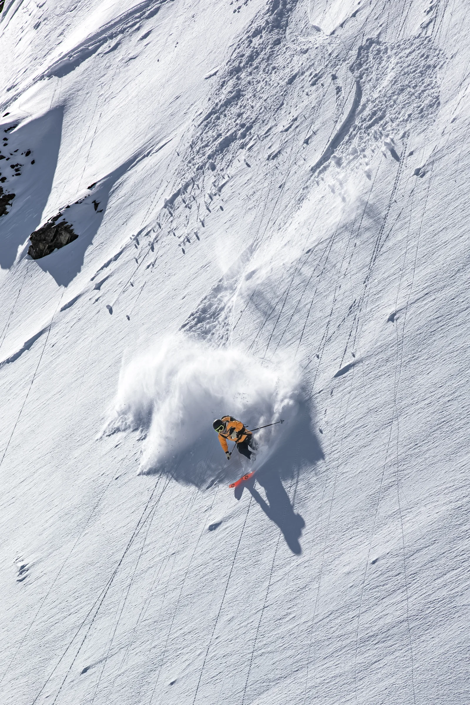 Michaela Casotti turns on a sunlit steep slope, throwing up a large cloud of snow as her long shadow stretches downhill.