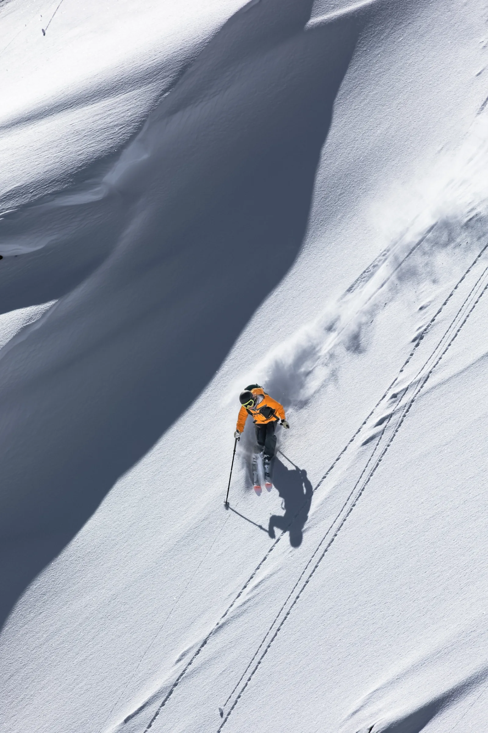 Michaela Casotti skis down a steep snowy slope in Lenzerheide in an orange jacket, trailing a plume of snow and casting a long shadow.