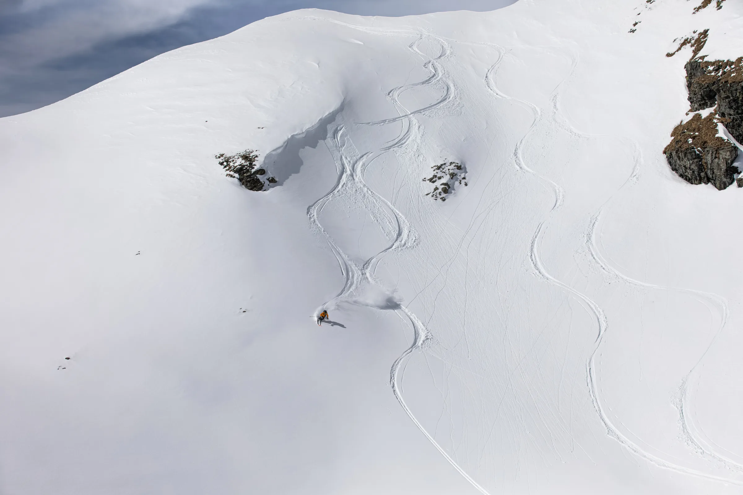 Michaela Casotti makes a turn through snow on a broad slope in Lenzerheide, below exposed rocks and numerous winding ski tracks.