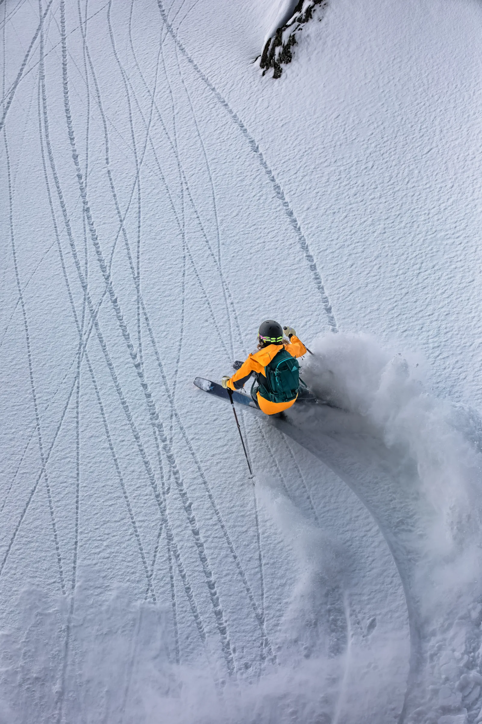 Seen from behind, Michaela Casotti wears an orange jacket and green backpack as she makes a deep turn on a tracked snowy slope, throwing up snow.