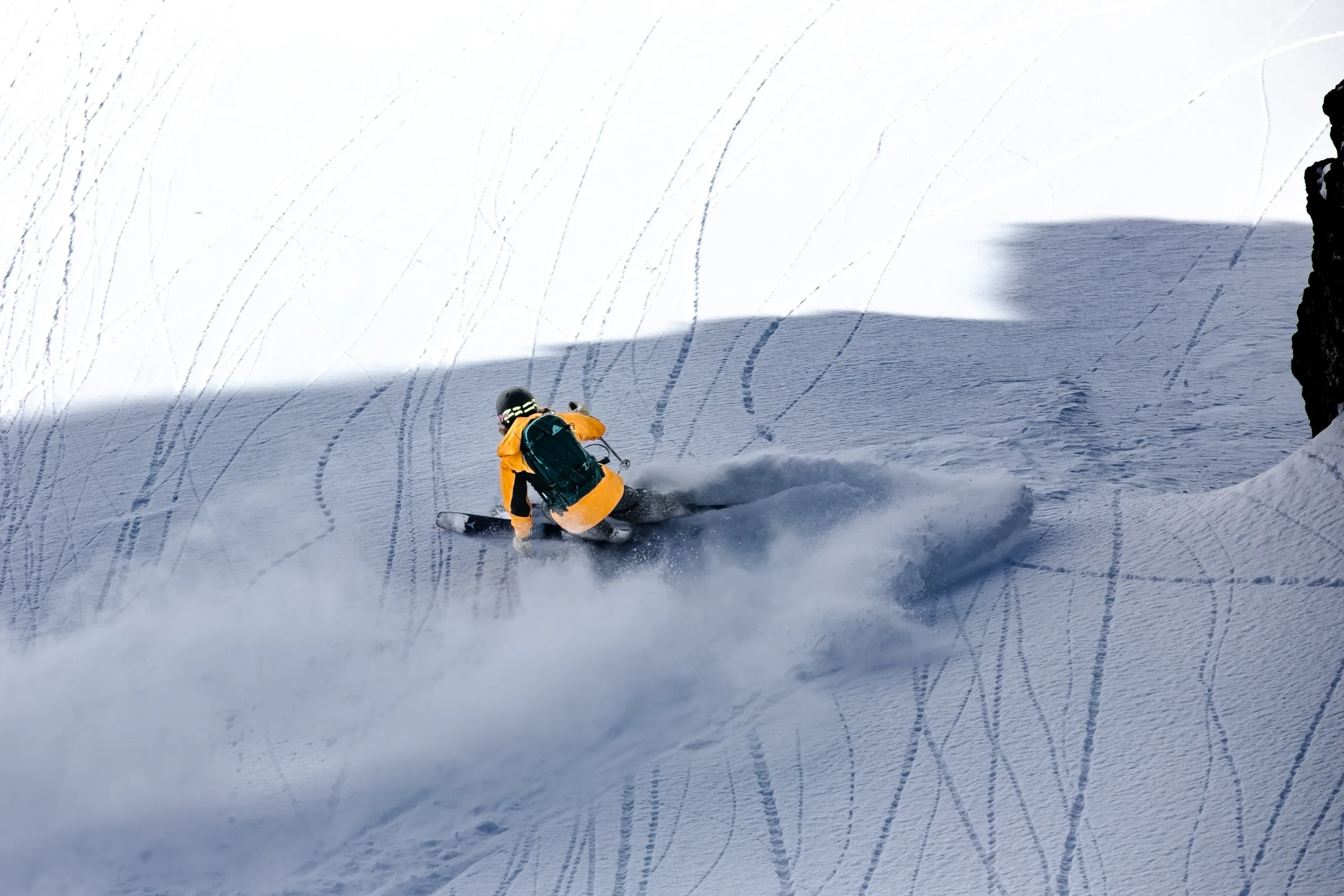 Michaela Casotti leans low into a ski turn on a shaded snowy slope beside a rock, leaving a broad cloud of snow behind her.