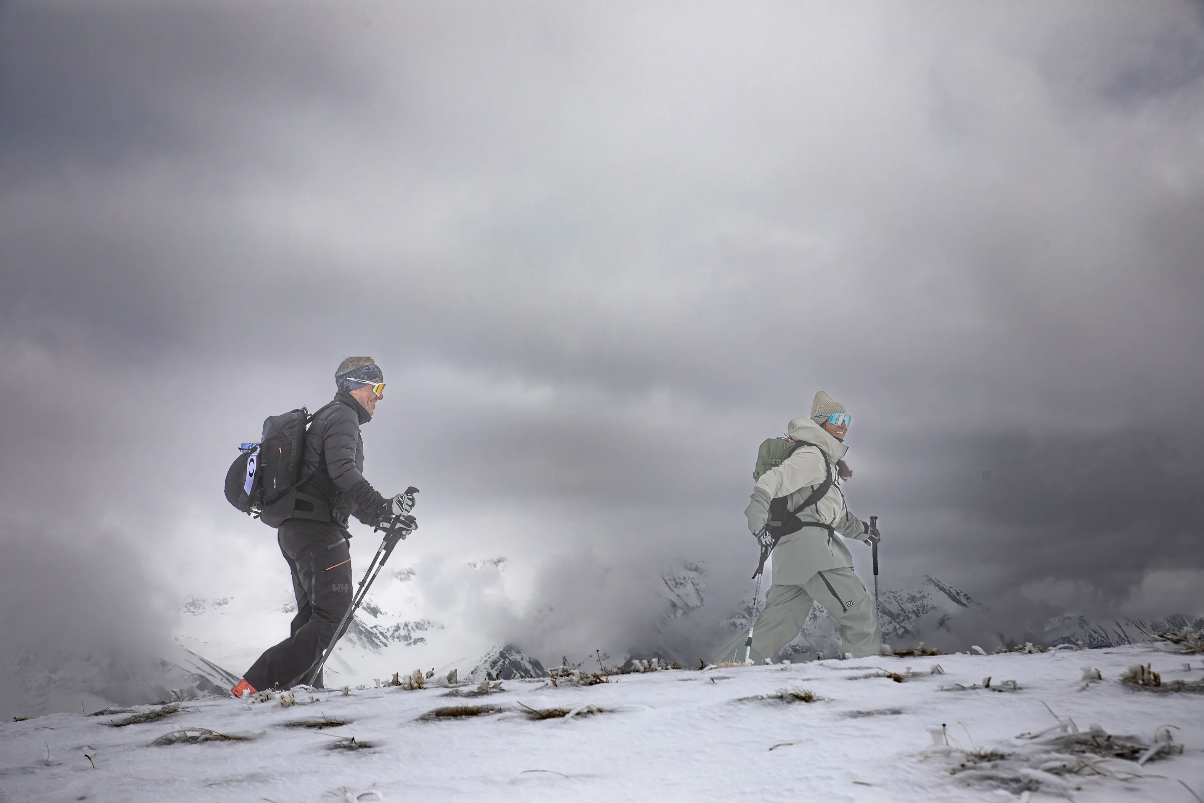 Zwei Personen gehen mit Rucksäcken und Stöcken über einen dünn verschneiten Hang bei Lenzerheide, hinter ihnen liegen Berggipfel in dichten Wolken.