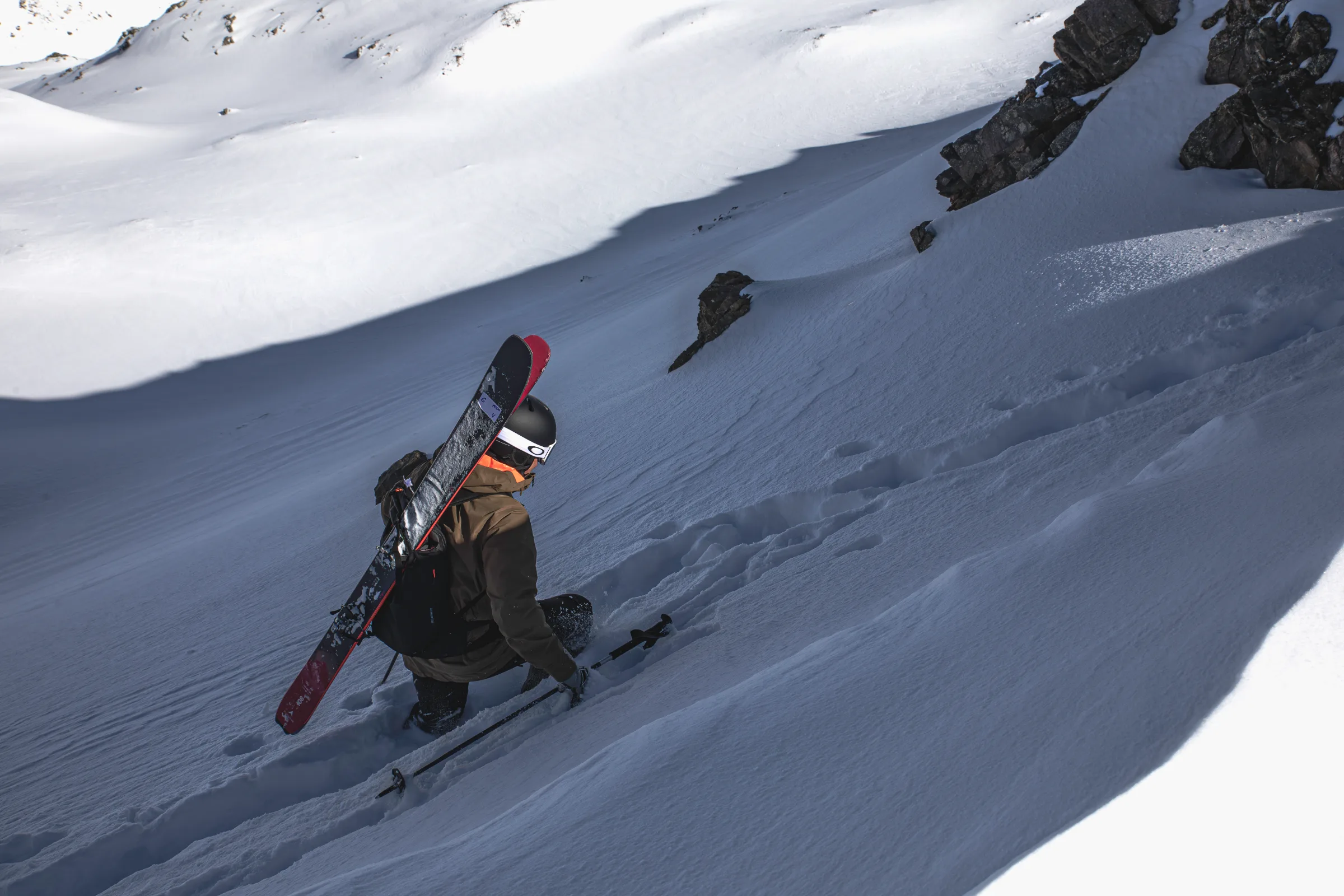 Mico Kehrle steigt mit Helm und diagonal am Rucksack befestigten Ski durch eine tiefe Fußspur einen schattigen Schneehang zwischen Felsen hinauf.