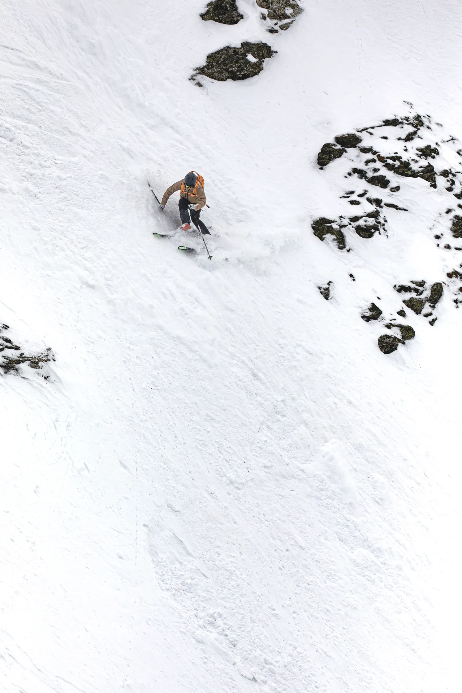 Mico Kehrle fährt mit brauner Jacke, Helm und orangefarbenem Rucksack eine steile, felsdurchsetzte Schneerinne in Lenzerheide hinab und wirbelt Schnee auf.