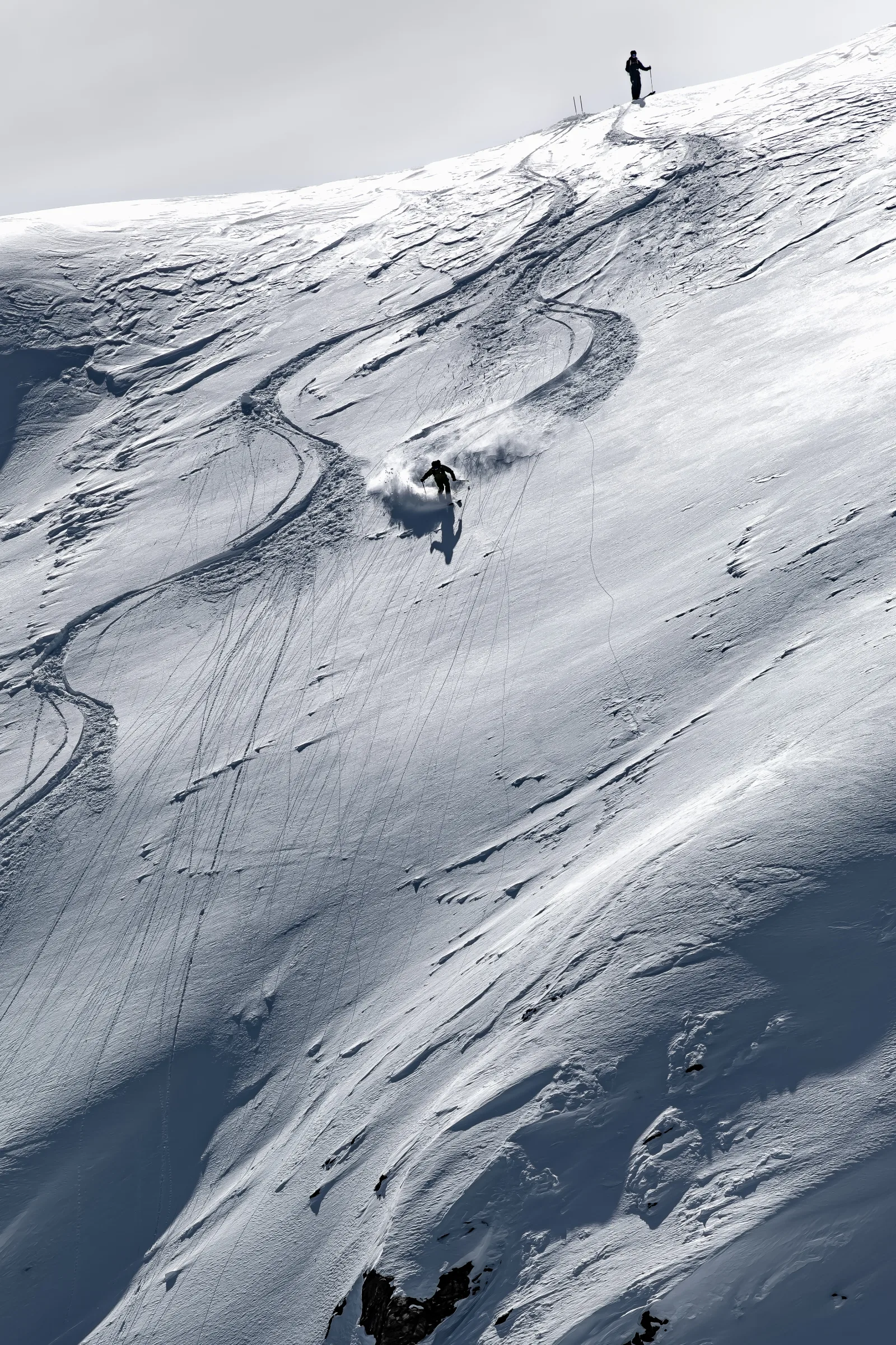 Mico Kehrle zieht einen Schwung auf einem steilen Schneehang mit geschwungenen Skispuren, während eine weitere Person oben am Grat steht.