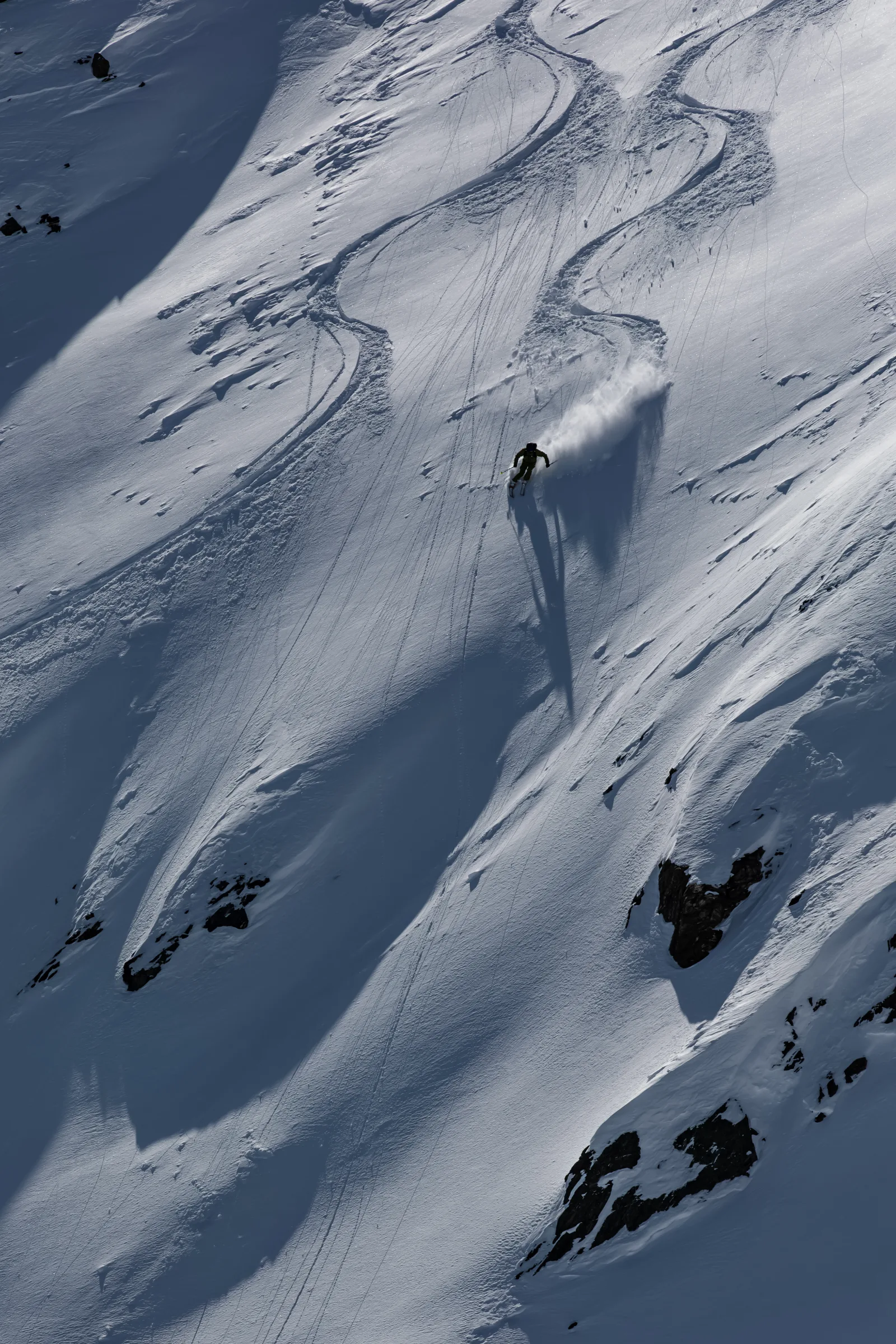 Mico Kehrle fährt einen steilen, felsdurchsetzten Schneehang hinab und zieht eine Pulverschneefahne hinter sich her; sein langer Schatten fällt talwärts.