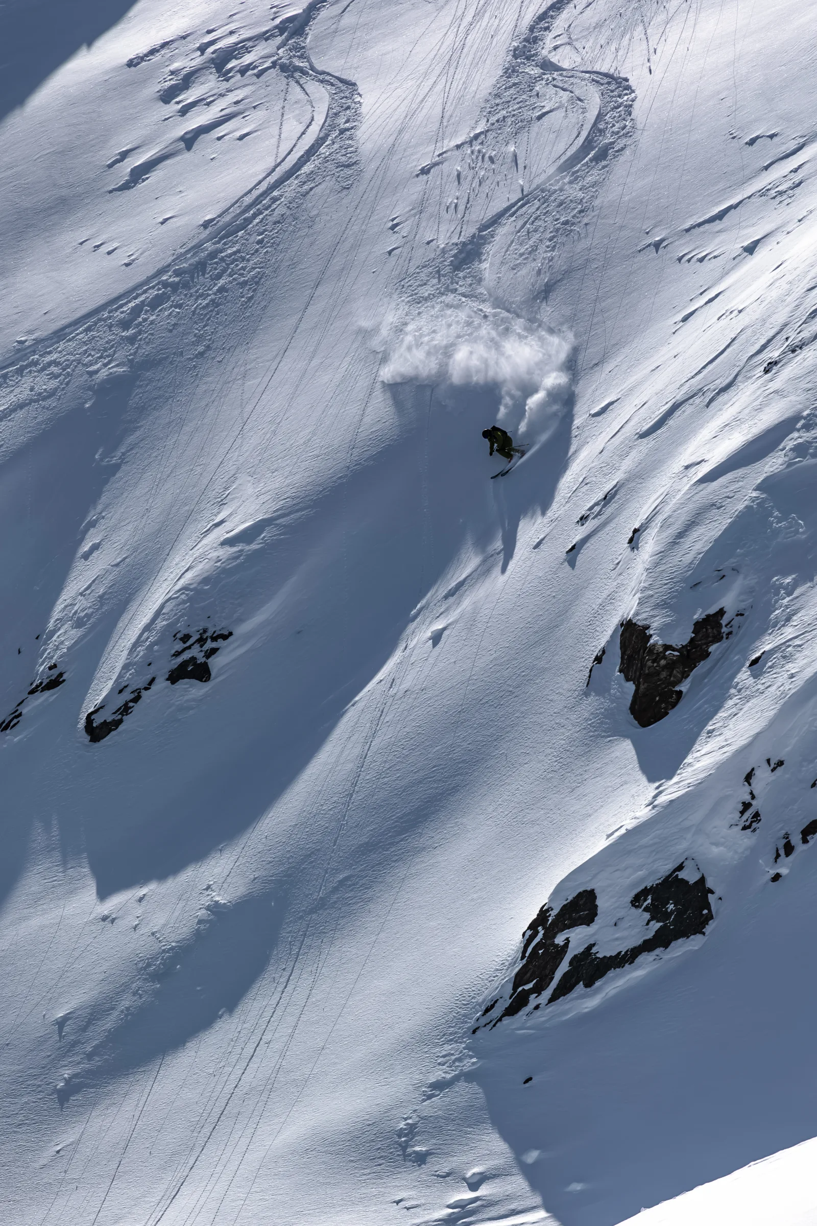 Mico Kehrle dreht auf einem steilen Schneehang neben freiliegenden Felsen in einen Schwung, über ihm hängt eine große aufgewirbelte Schneewolke.