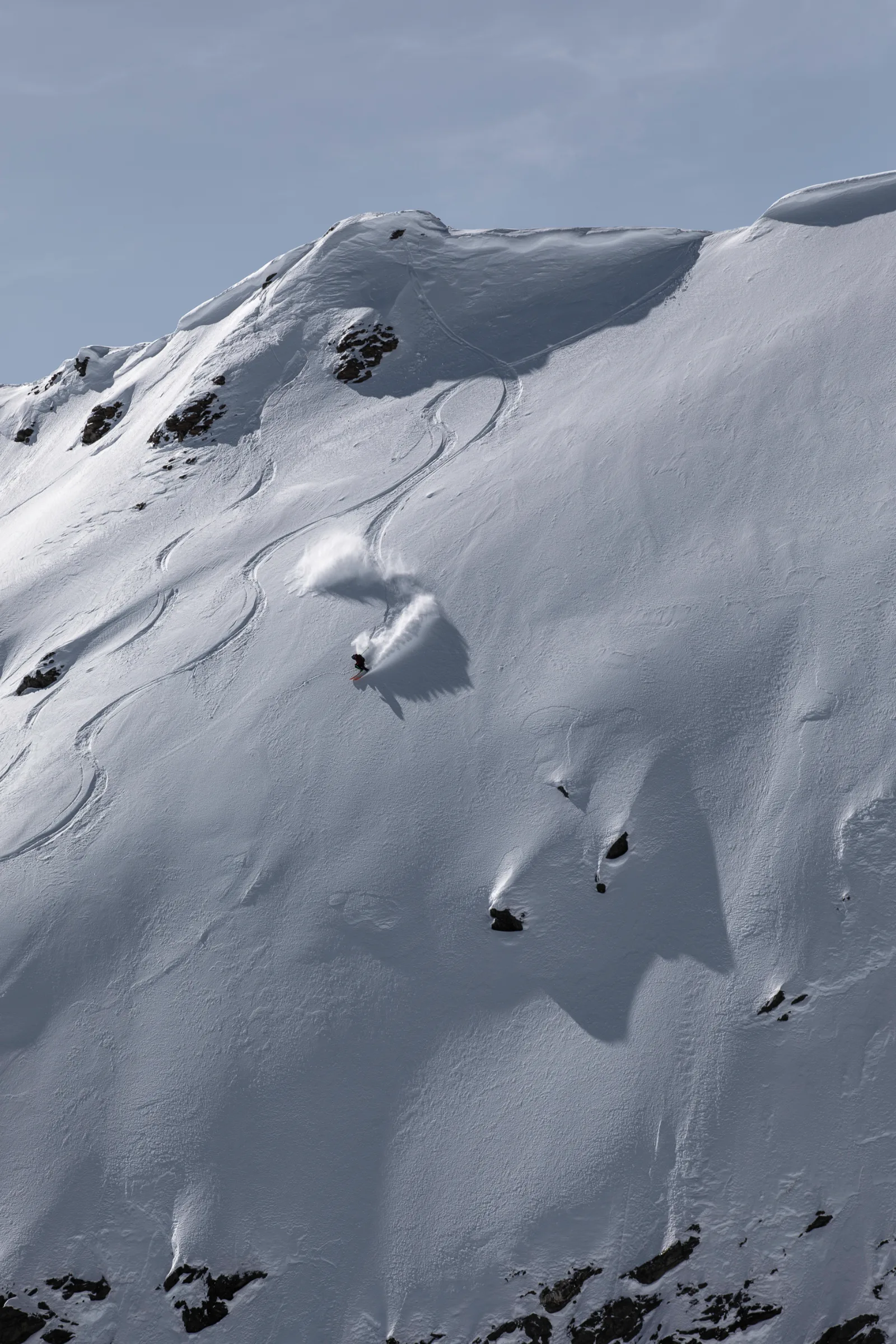Mike Perl zieht einen Schwung durch einen steilen Pulverschneehang in Lenzerheide und wirbelt zwischen einzelnen Felsen eine große Schneewolke auf.
