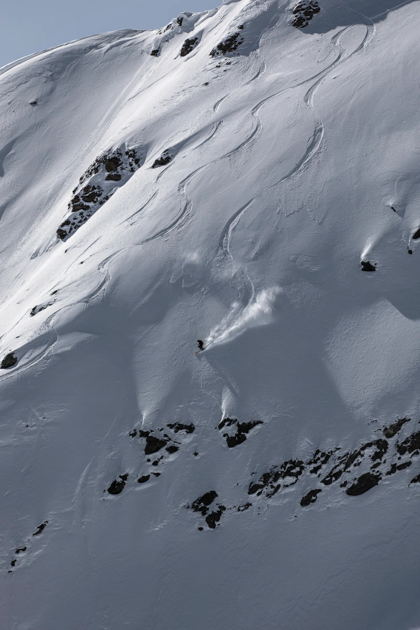 Mike Perl fährt mit einer Schneefahne hinter sich einen steilen Hang hinab, oberhalb dunkler Felsbänder und unter geschwungenen Skispuren.