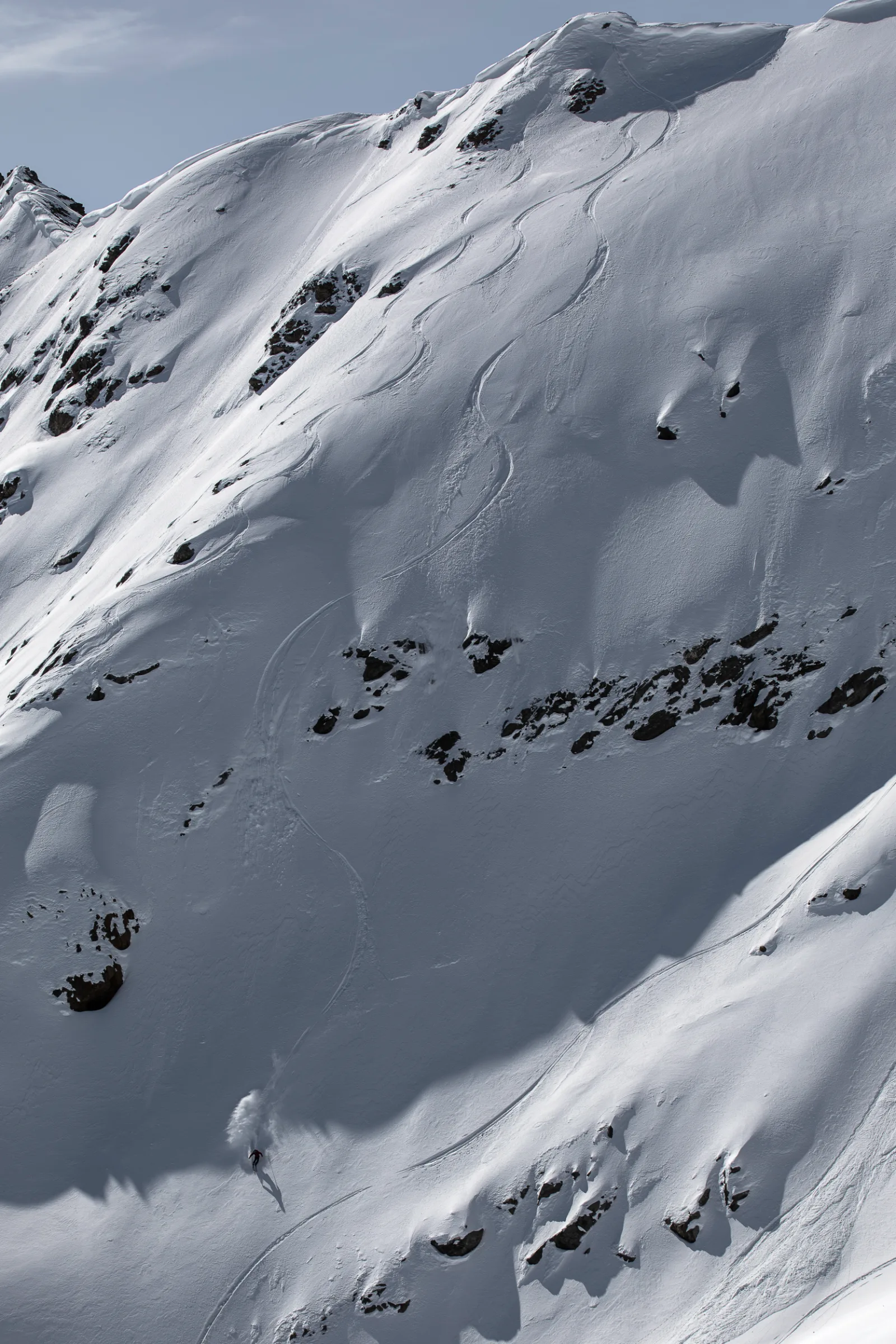 Mike Perl fährt am unteren Ende eines weiten Schneehangs in Lenzerheide durch den Pulverschnee, hoch über ihm verlaufen Skispuren zwischen Felsbändern.