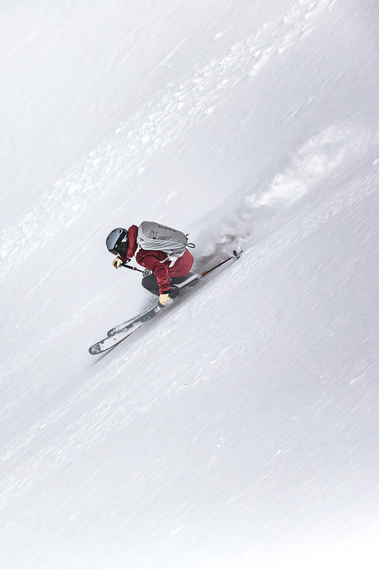 Von oben gesehen fährt Mike Perl tief geduckt über einen steilen Schneehang und wirbelt hinter seinen Ski Schnee auf; sein heller Rucksack hebt sich von der roten Jacke ab.