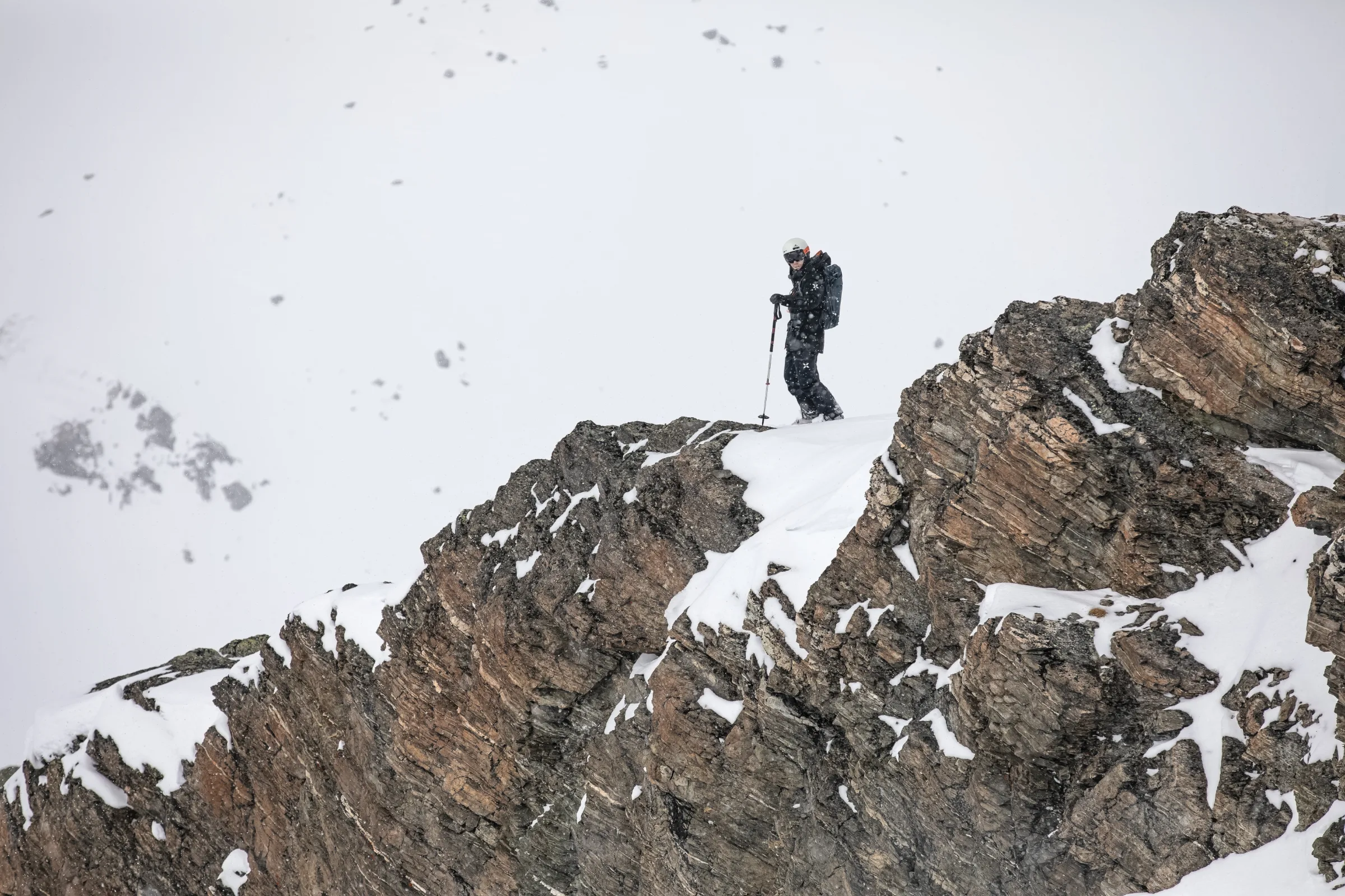 Mike Perl steht mit Helm, Skistöcken und Rucksack auf einem schmalen Schneestreifen zwischen zerklüfteten Felsen im Schneefall.