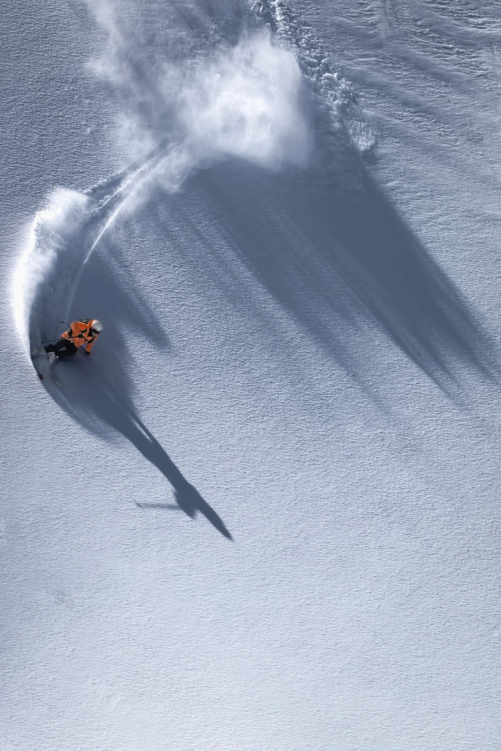 Von oben gesehen legt sich Mike Perl in einen Schwung auf einem offenen Schneehang, begleitet von aufsprühendem Schnee und einem langen Schatten.