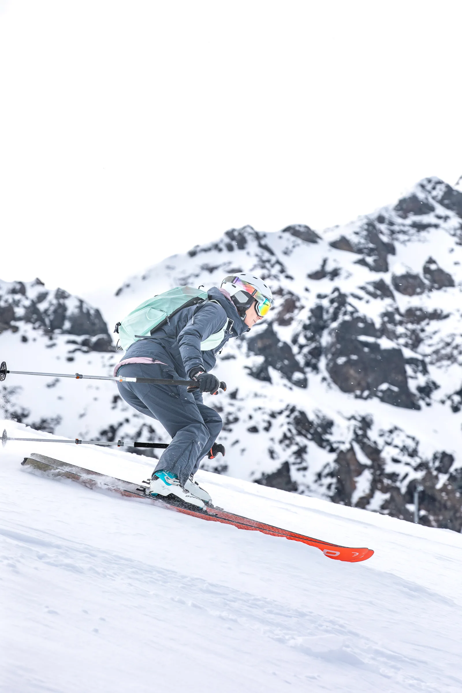 Petra Rapp fährt mit mintgrünem Rucksack und gebeugten Knien einen Schneehang in Lenzerheide hinab, vor schneebedeckten Felsgipfeln.