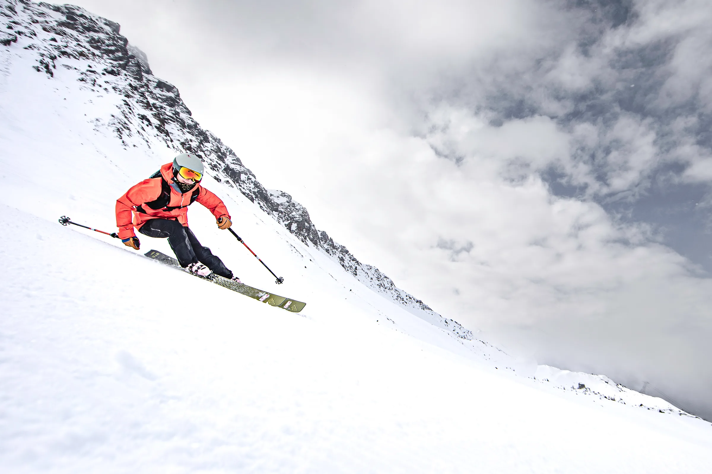 Pia Vogt skis with bent knees down a steep snowy slope in Lenzerheide, wearing a red jacket, with a rocky ridge and clouds behind her.
