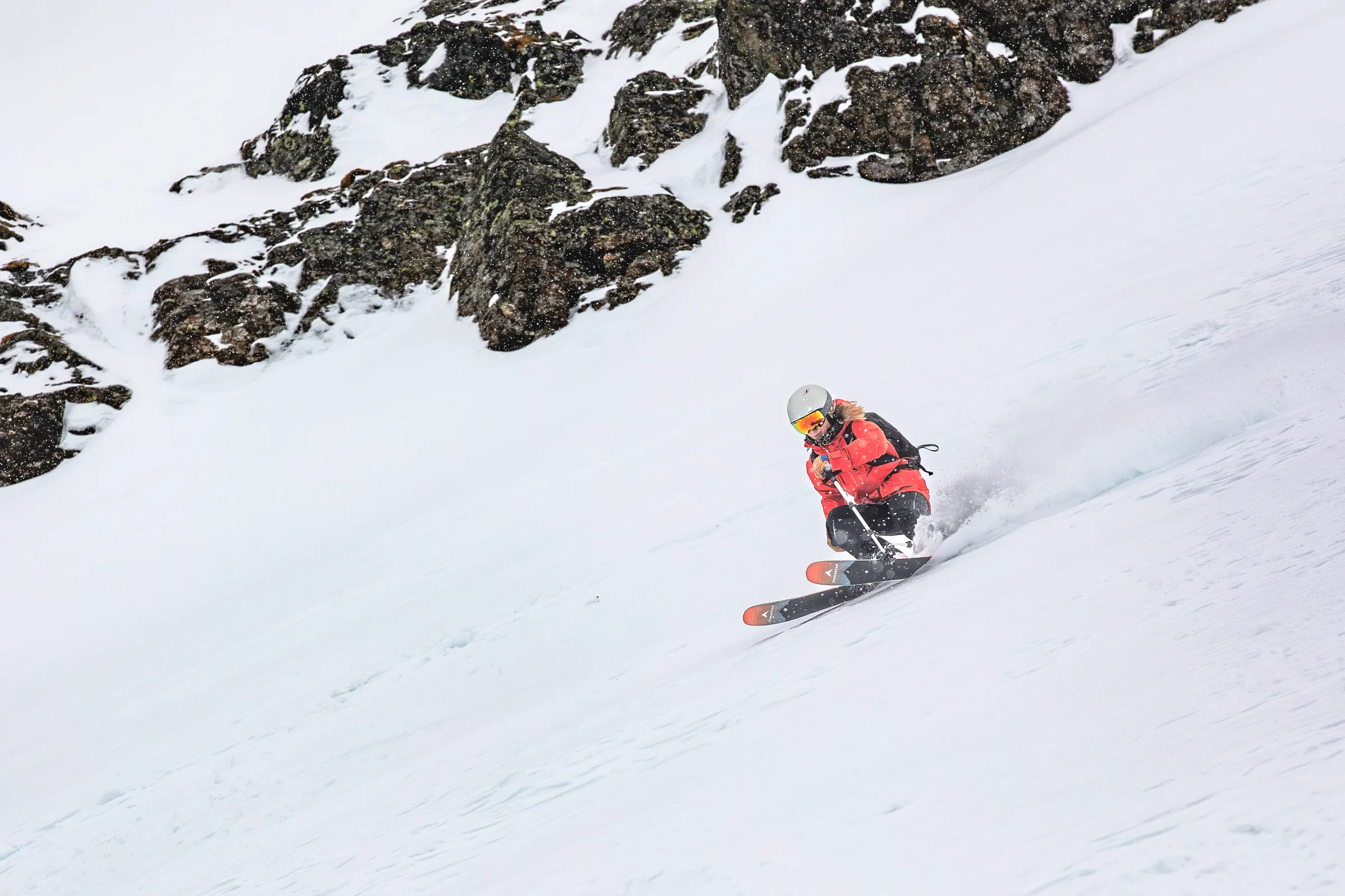 Pia Vogt makes a low, crouched turn below snow-covered rocks, sending snow spraying behind her skis.