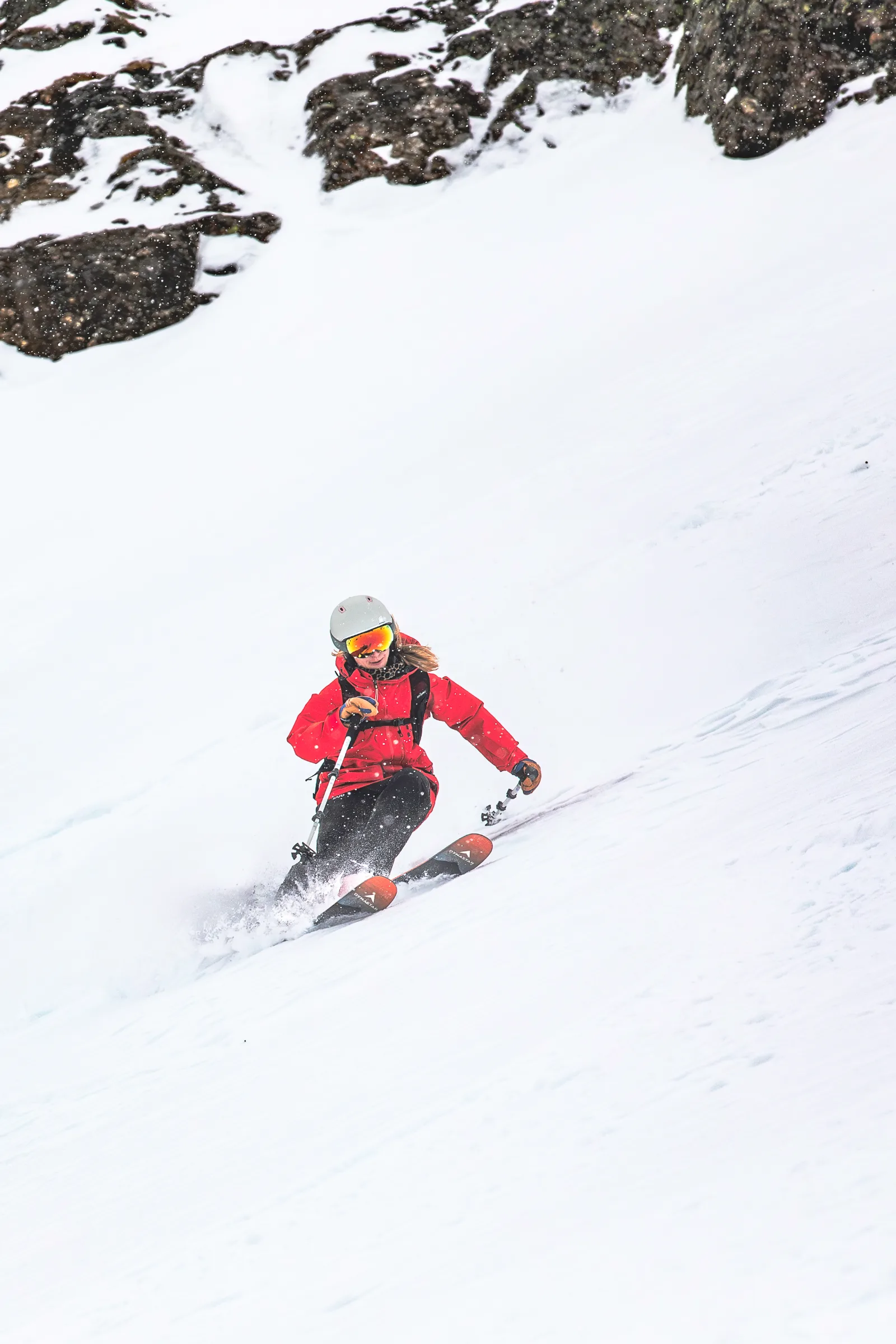 Wearing a red jacket, grey helmet and mirrored goggles, Pia Vogt turns down a snowy slope beneath dark rocks.