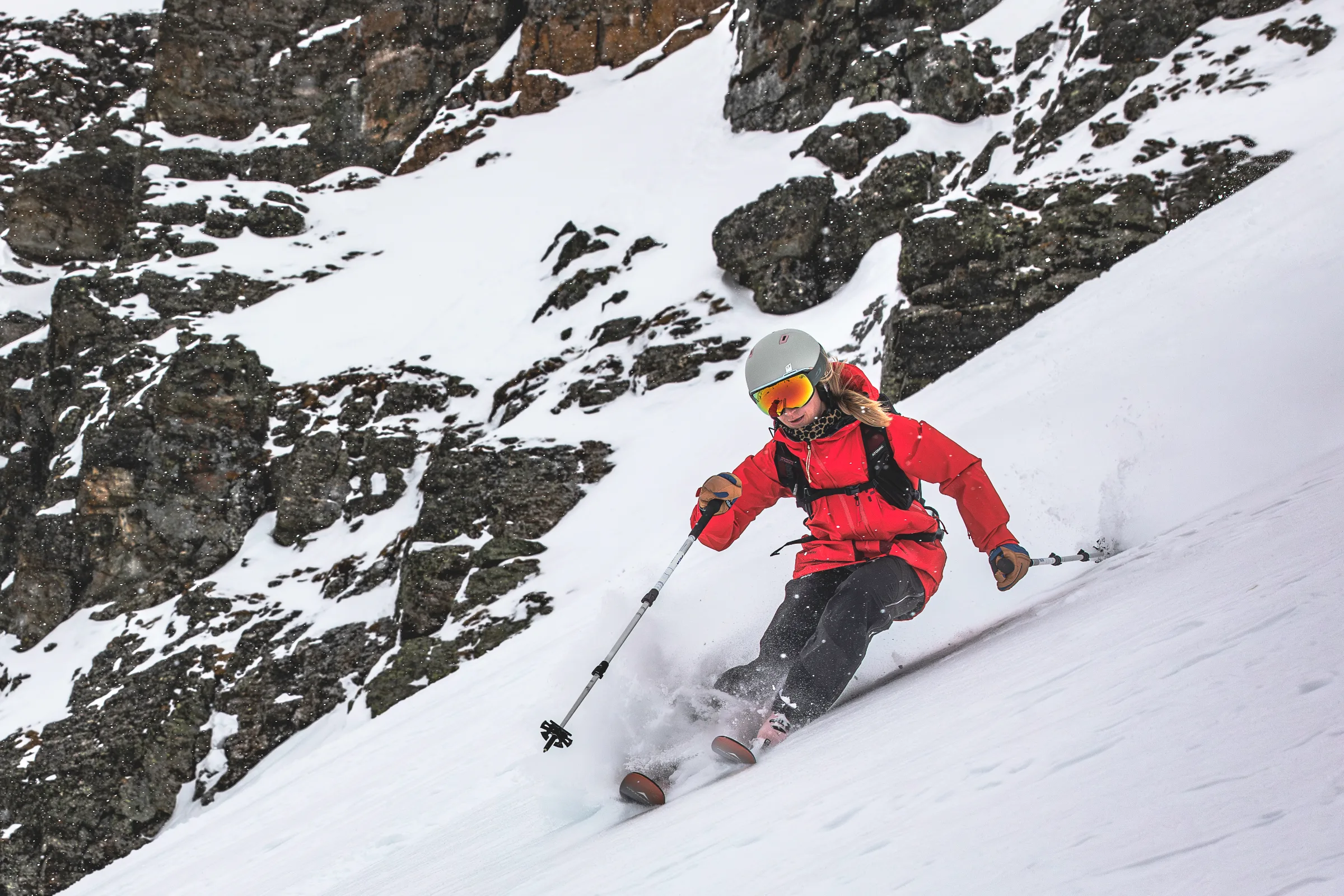 Pia Vogt skis past a snow-covered rock face with parallel skis and poles held apart, snow spraying around her boots.