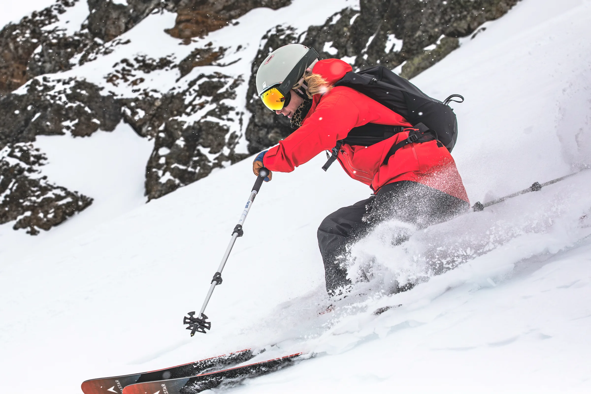 In a close side view, Pia Vogt skis through a turn in a red jacket and black backpack, throwing up snow against a rocky backdrop.