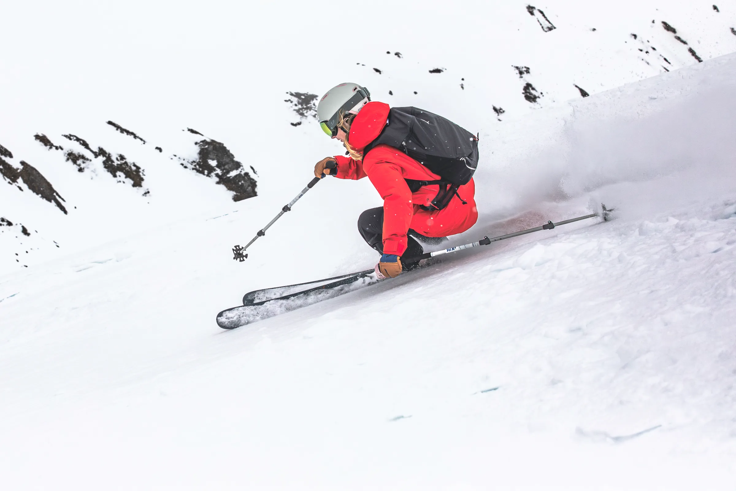 Seen from behind and to the side, Pia Vogt skis in a deep crouch across a steep snowy slope, trailing a plume of snow.
