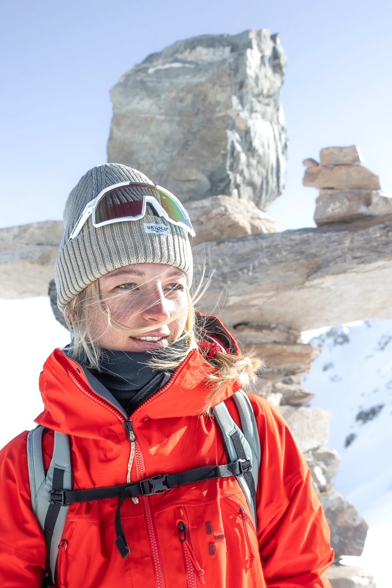 Pia Vogt smiles and looks to the side, wearing a grey knitted hat with sunglasses perched on it, beside boulders and snow under a blue sky.