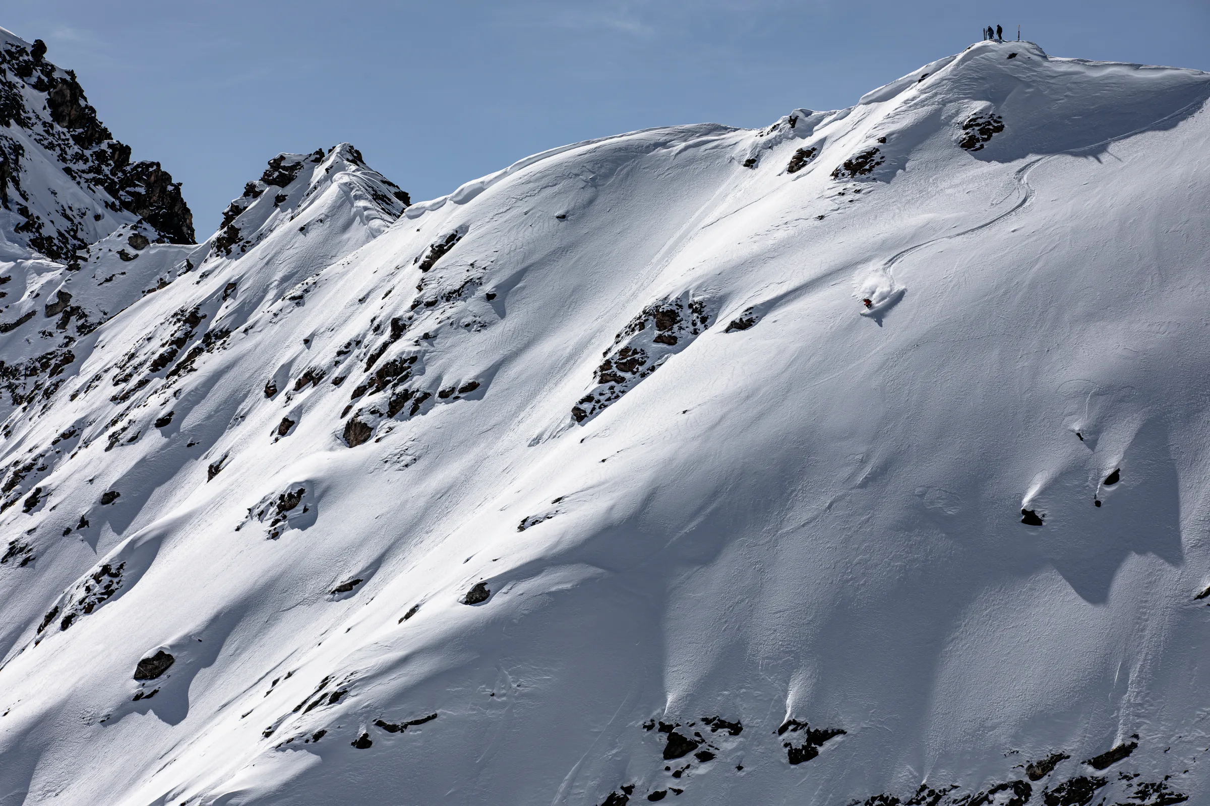 Pia Vogt makes a turn with a plume of snow on a steep, rocky slope in Lenzerheide, while several people stand on the ridge above.
