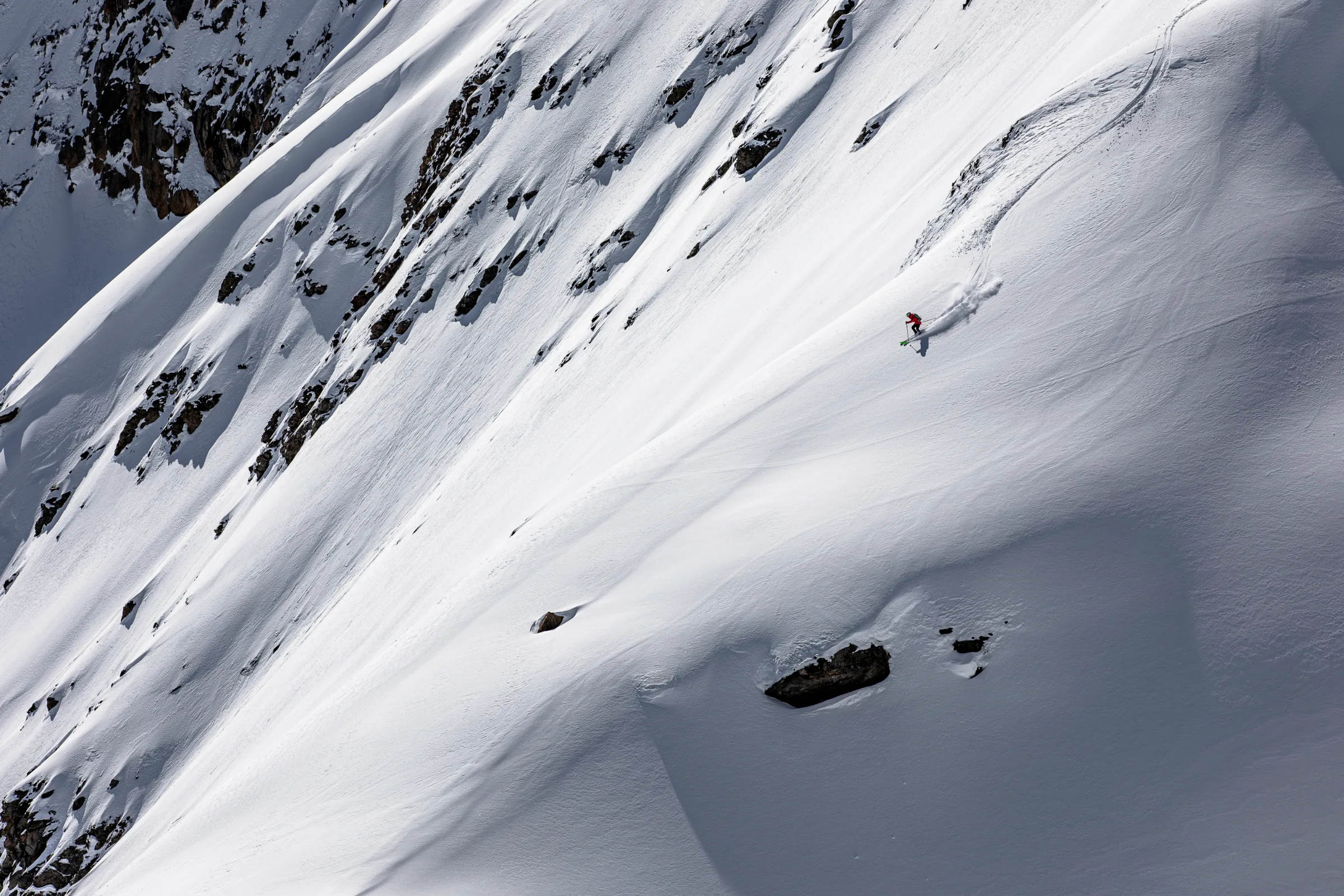 Wearing a red jacket, Pia Vogt skis on green skis across an open snowy slope below steep rocky faces, throwing up a trail of snow.